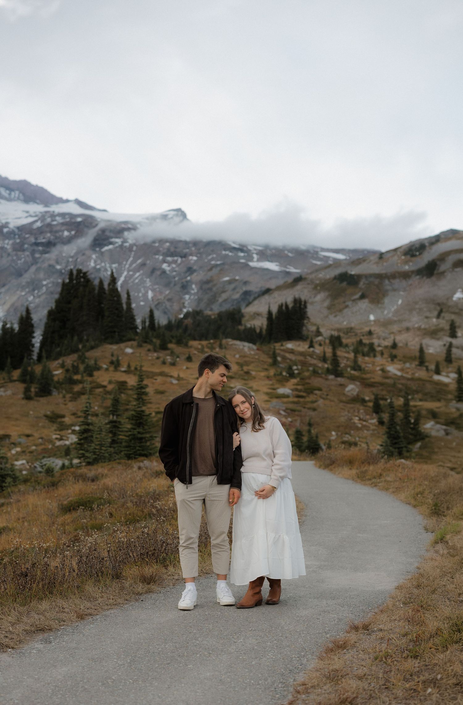 Couple standing on a mountain path with snowy peaks and evergreen trees in the background on a cloudy day.