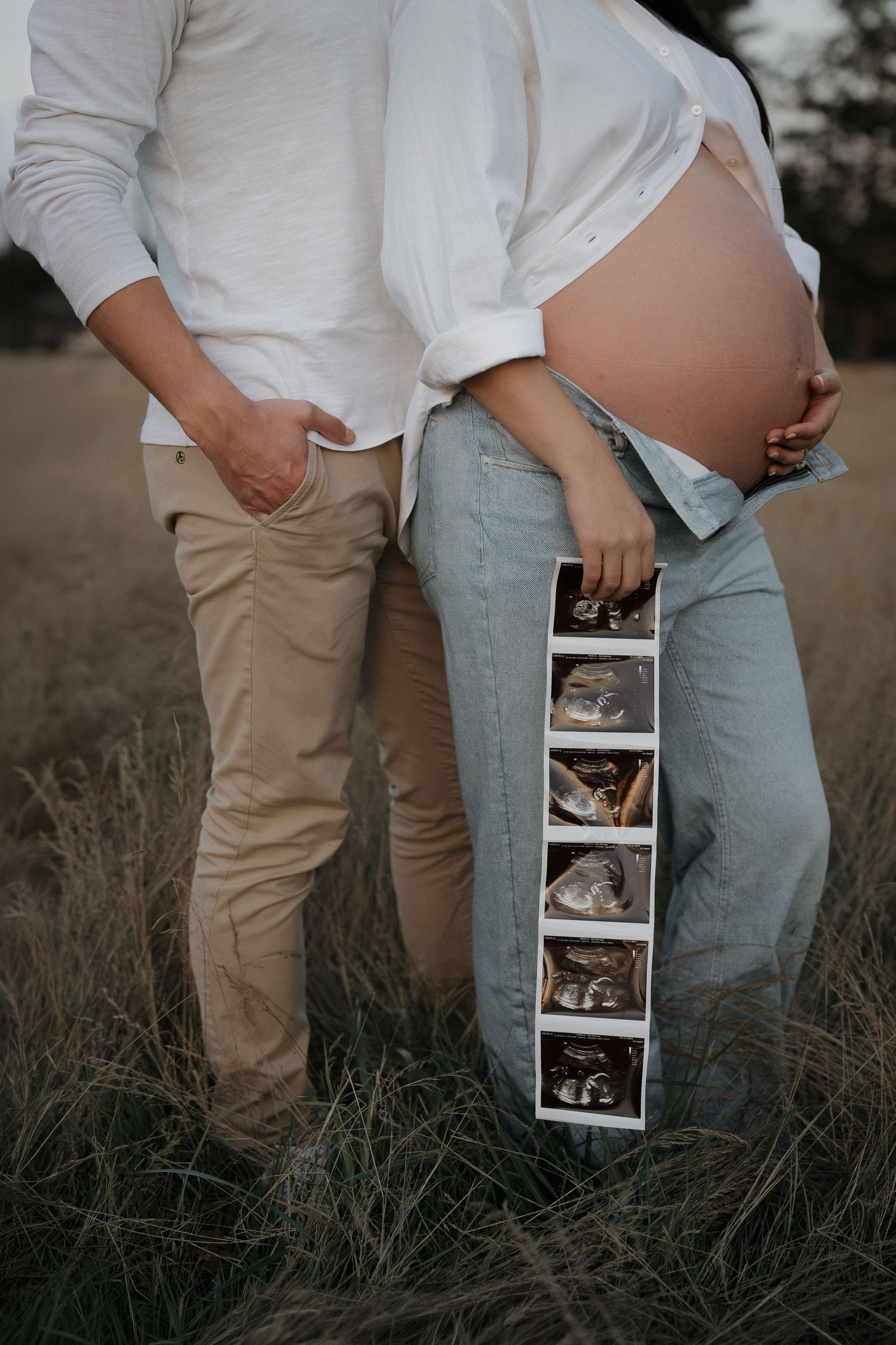 Expecting couple standing in a grassy field, the pregnant woman's bare belly showing as she holds a strip of ultrasound photos.