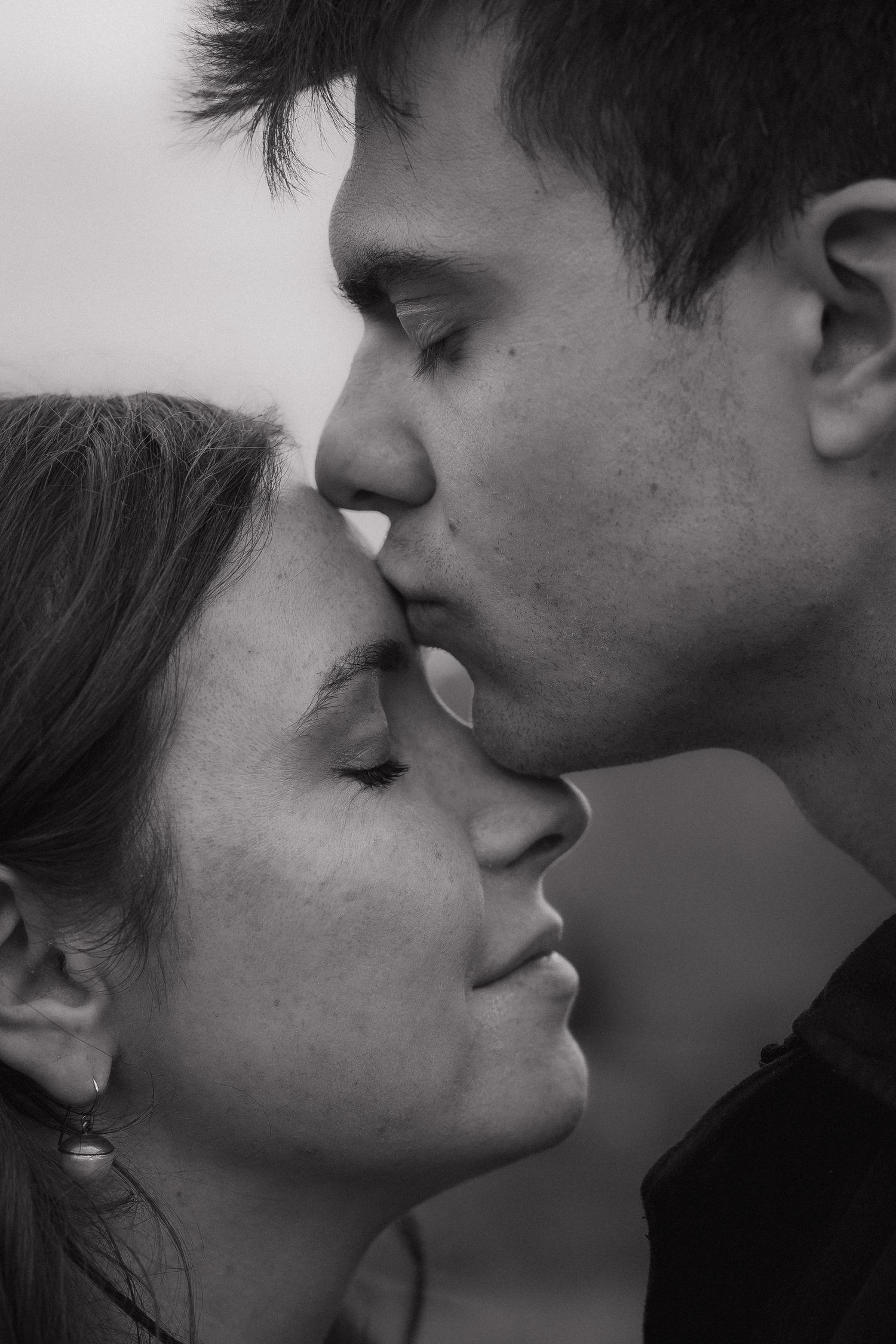 Black and white photo of a young couple sharing a tender forehead kiss with eyes closed.