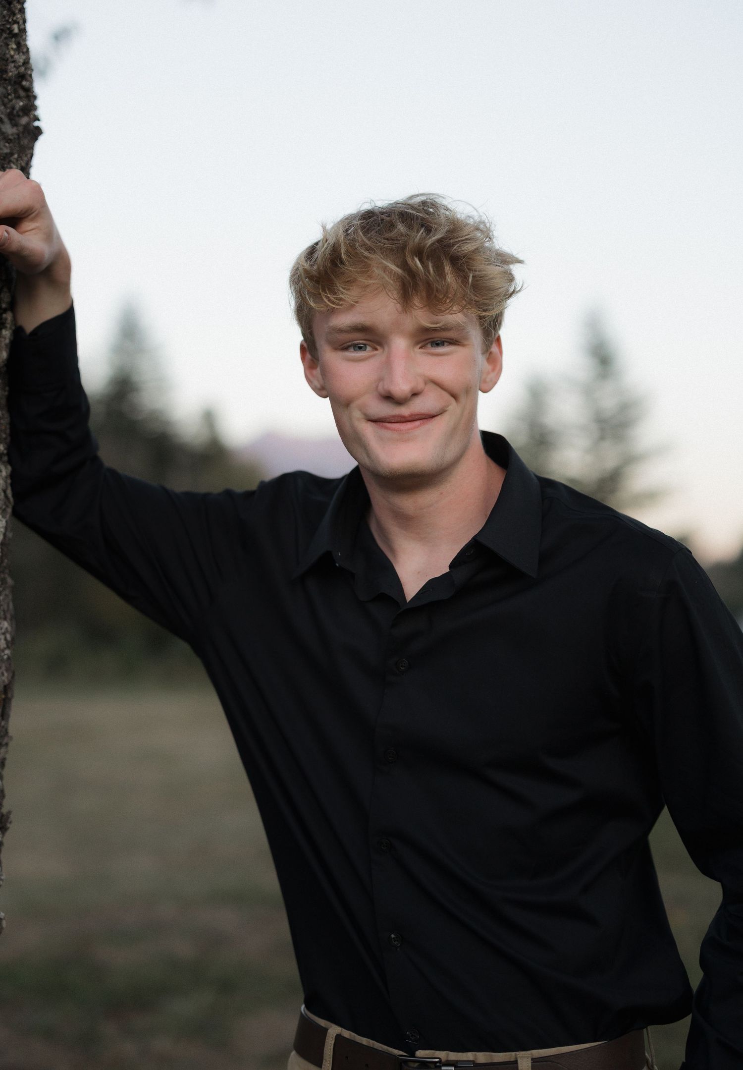 Young blond man in black shirt leaning against a tree, smiling outdoors at dusk.