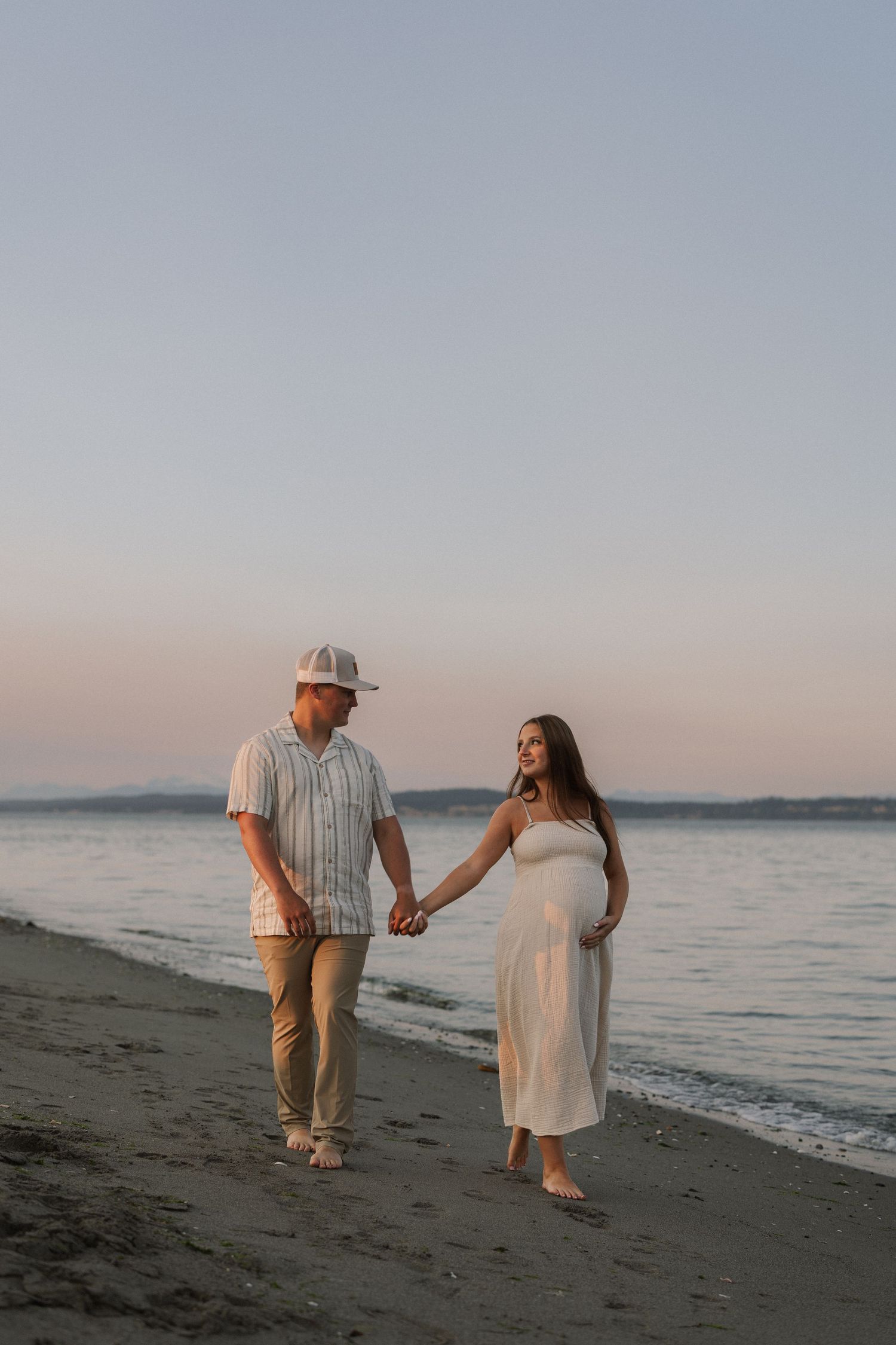 Pregnant couple holding hands walking on beach at sunset, woman in white dress, man in striped shirt and cap.