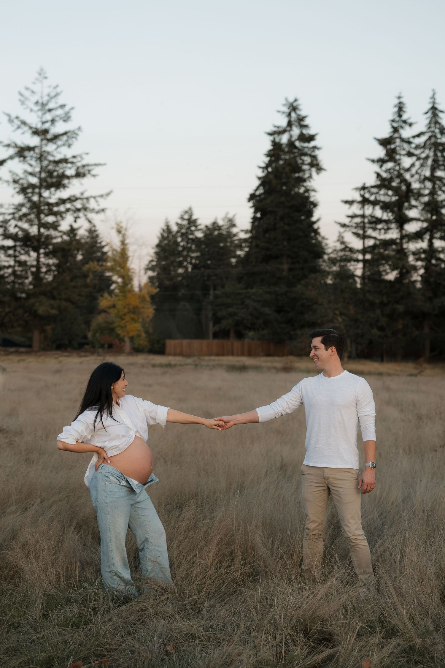 Expecting couple holding hands during an outdoor maternity photoshoot in a grassy field with tall evergreen trees at golden hour.