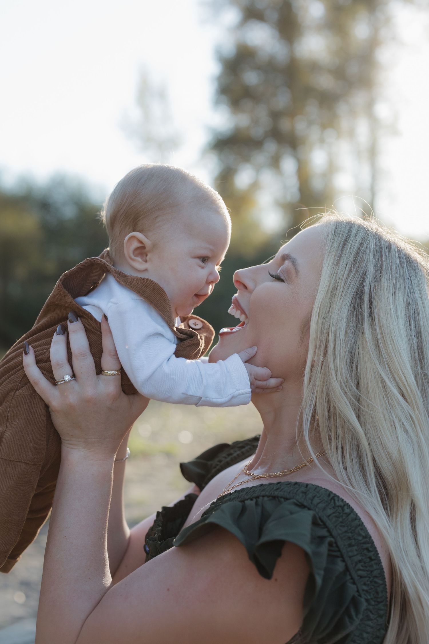 A joyful blonde mother lifts her smiling baby outdoors in warm sunlight, sharing a tender moment of love and laughter.