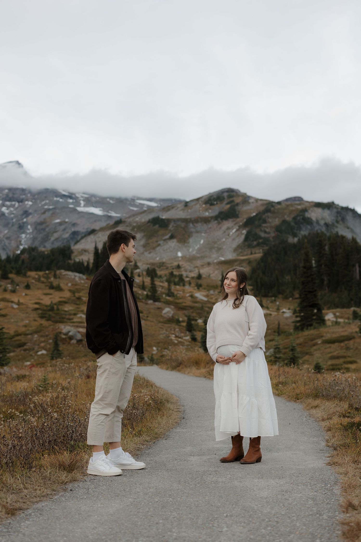 Couple standing on a mountain path with misty peaks and autumn foliage in the background.
