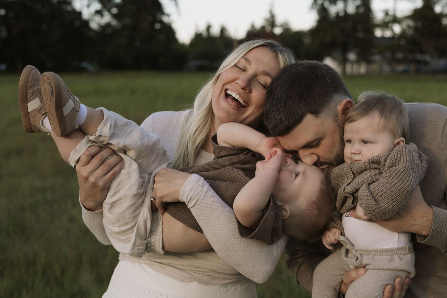 Happy family with laughing mom, dad kissing toddler, and baby outdoors in green field during golden hour photo session.