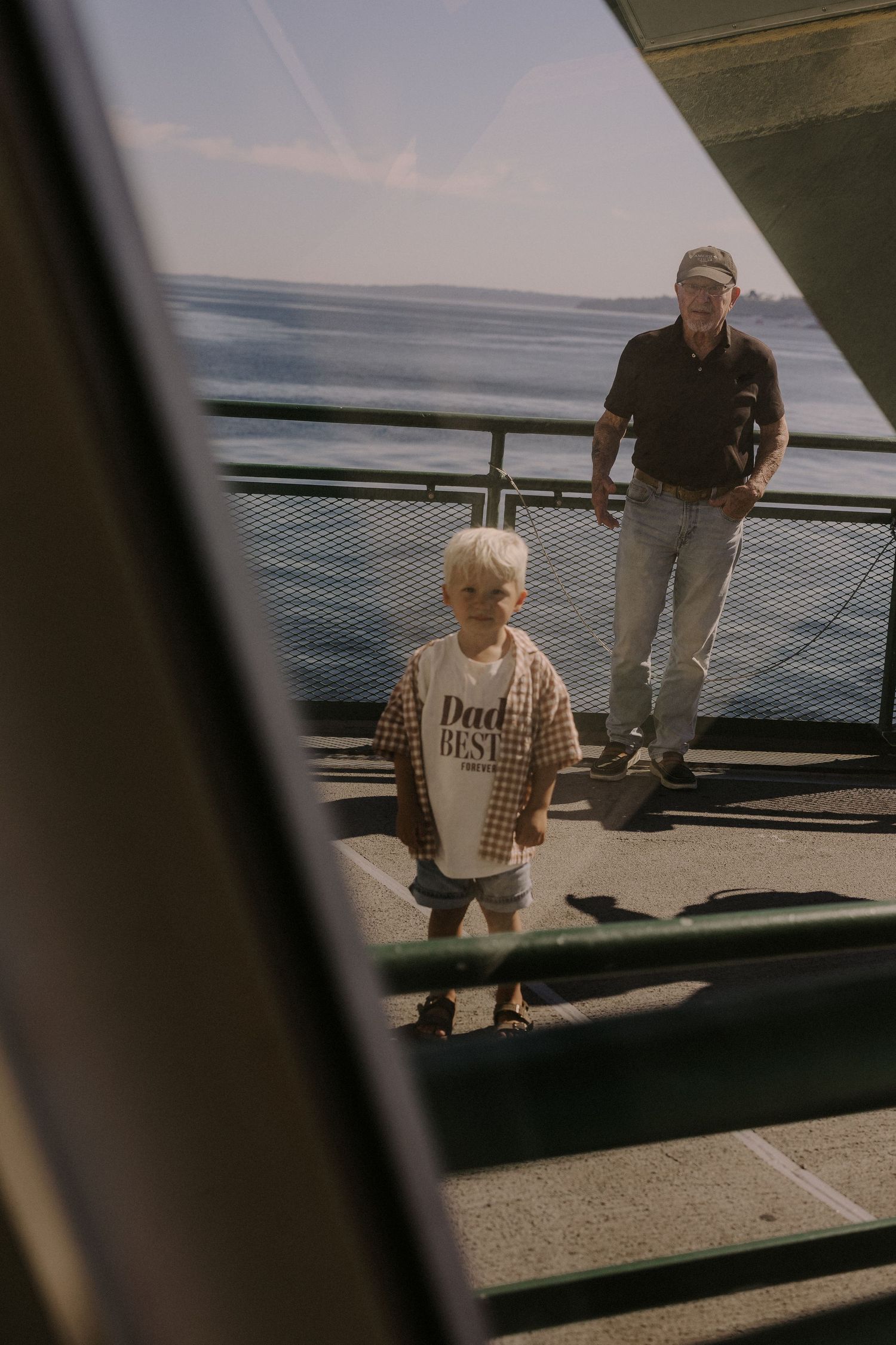 A young child in a Dad shirt stands on a sunny ferry deck with an older man behind him.