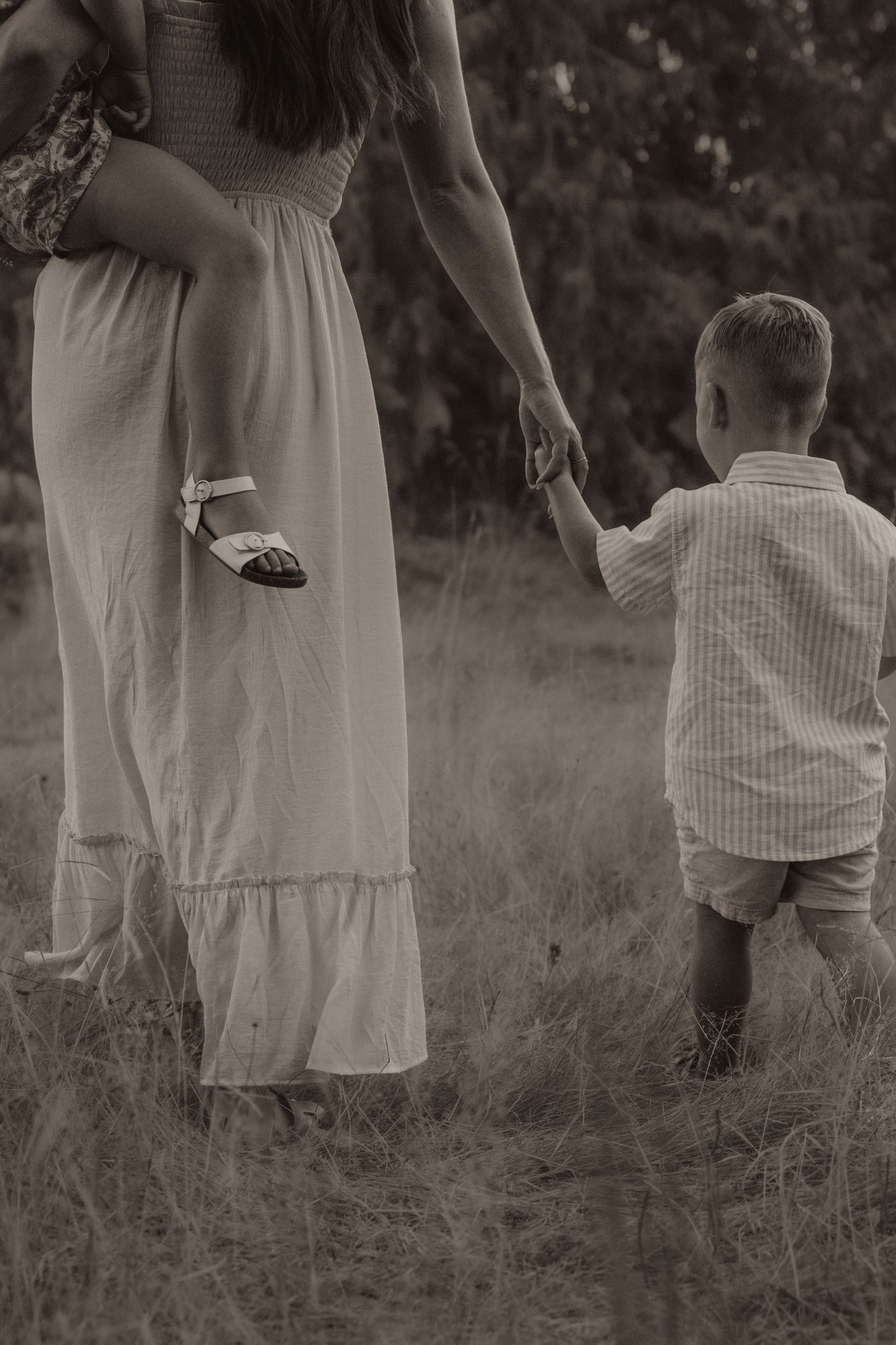 Sepia-toned photo of a mother holding a toddler while walking hand-in-hand with a young boy through a grassy field.
