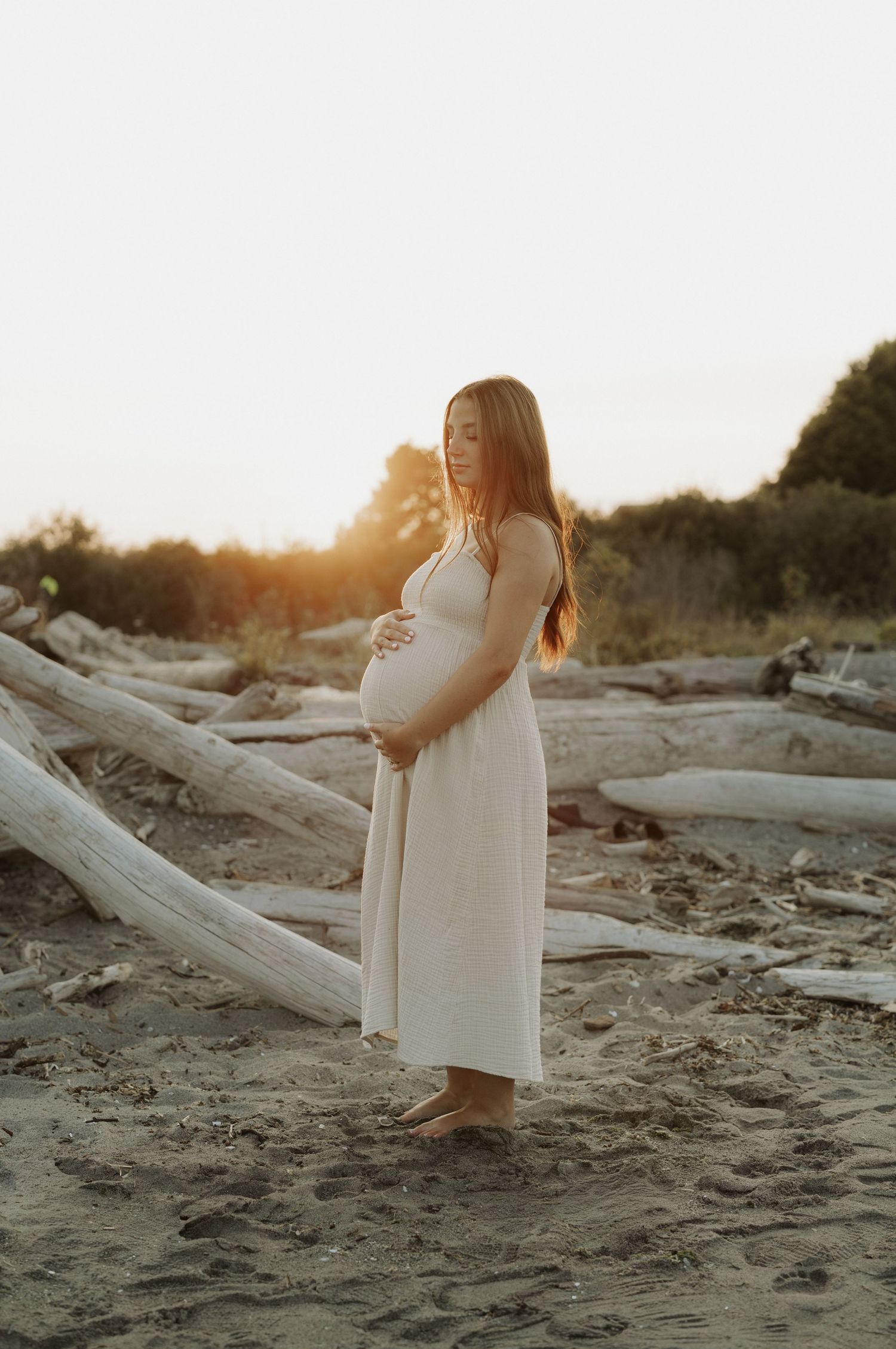 Pregnant woman in white dress cradling baby bump on sandy beach at golden sunset with driftwood.