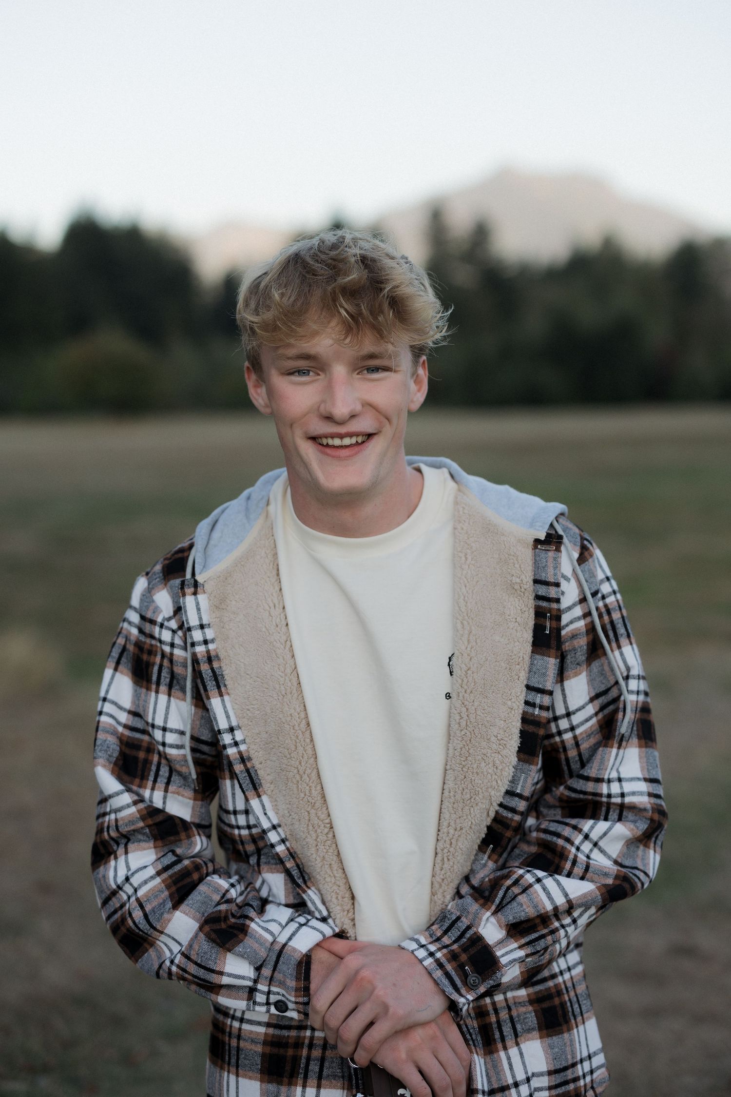 Smiling young man in plaid sherpa jacket and white shirt standing in an open field with mountains in the background.