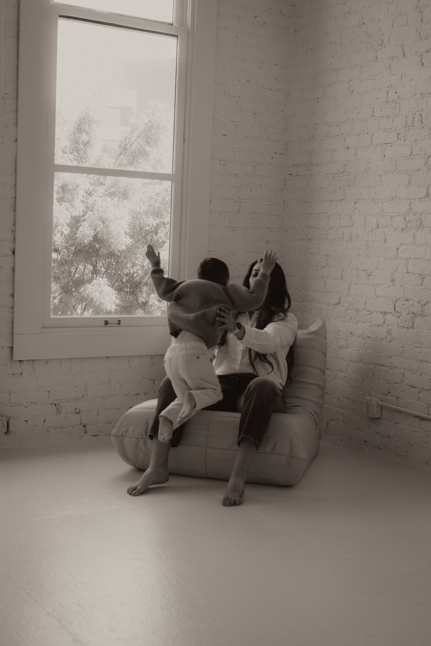 A mother and child playfully interact on a bean bag chair near a large window in a bright white brick room.