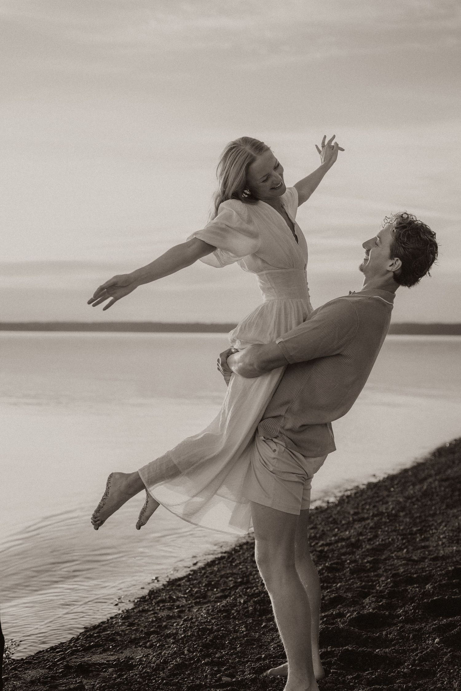 A man lifts a woman in a white dress on a rocky beach shore in a romantic black and white photo.