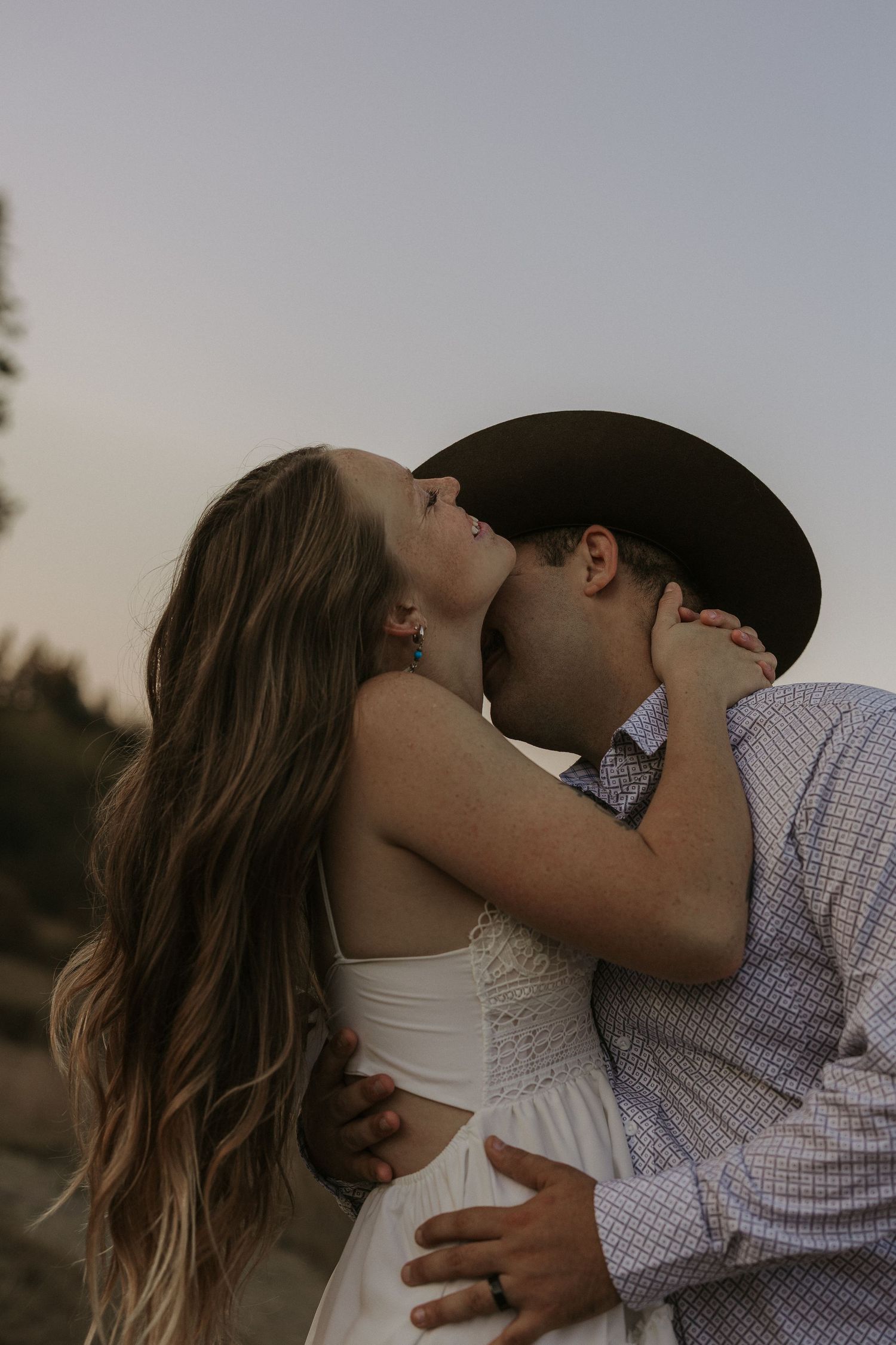 Romantic couple sharing a kiss outdoors at dusk, man wearing a dark cowboy hat.