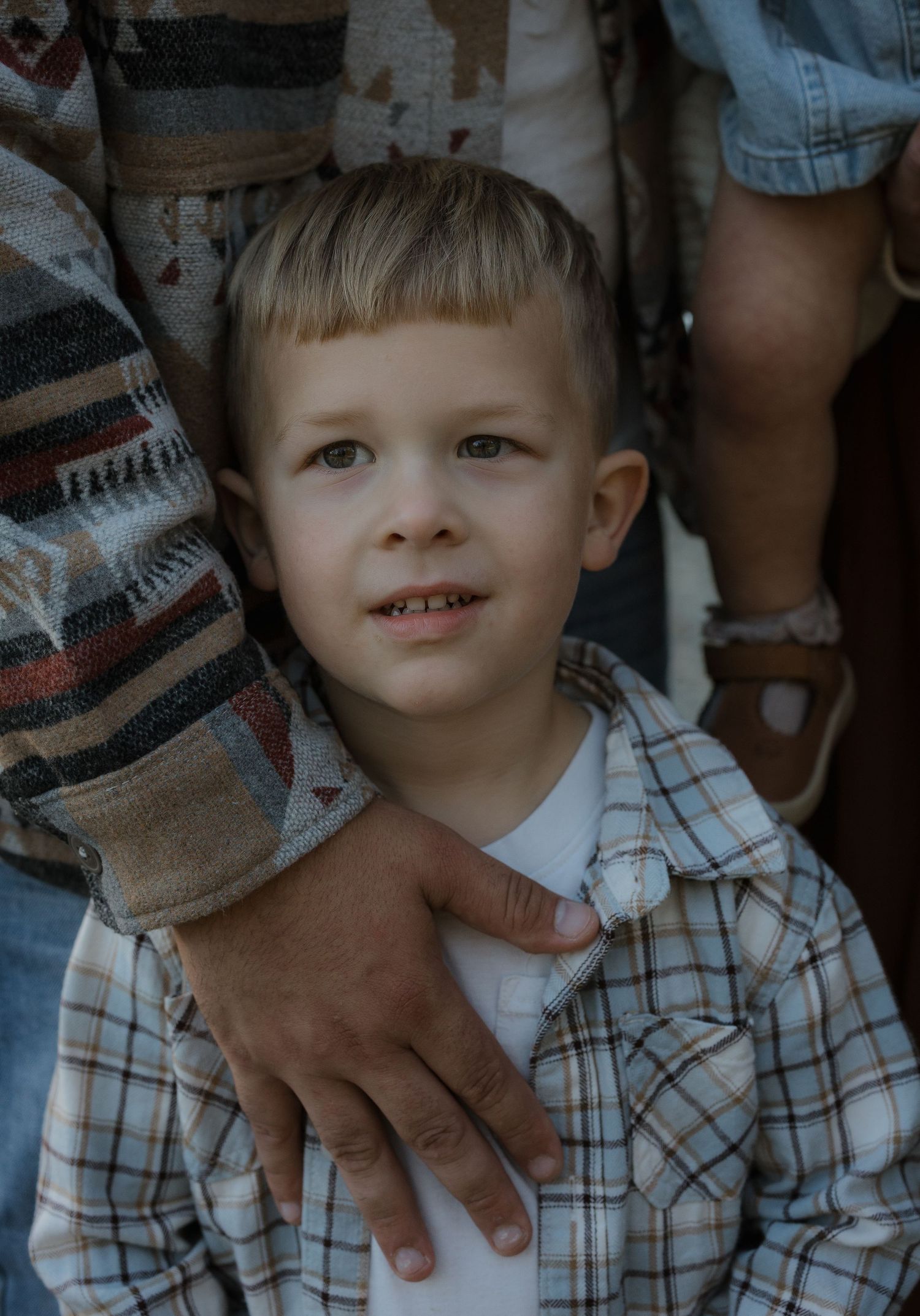 A young blonde boy in a plaid shirt looks up while an adult's hand rests on his shoulder.