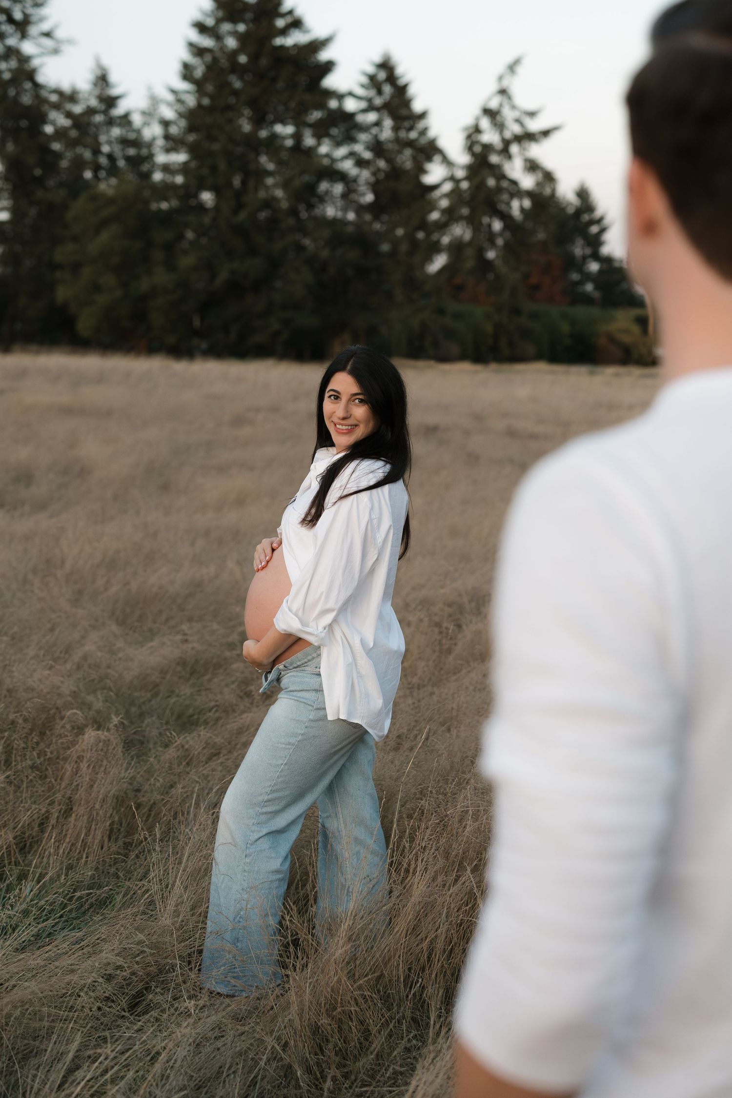 Smiling pregnant woman in a white shirt and jeans cradles her baby bump in a grassy field while a man stands blurred in the foreground.
