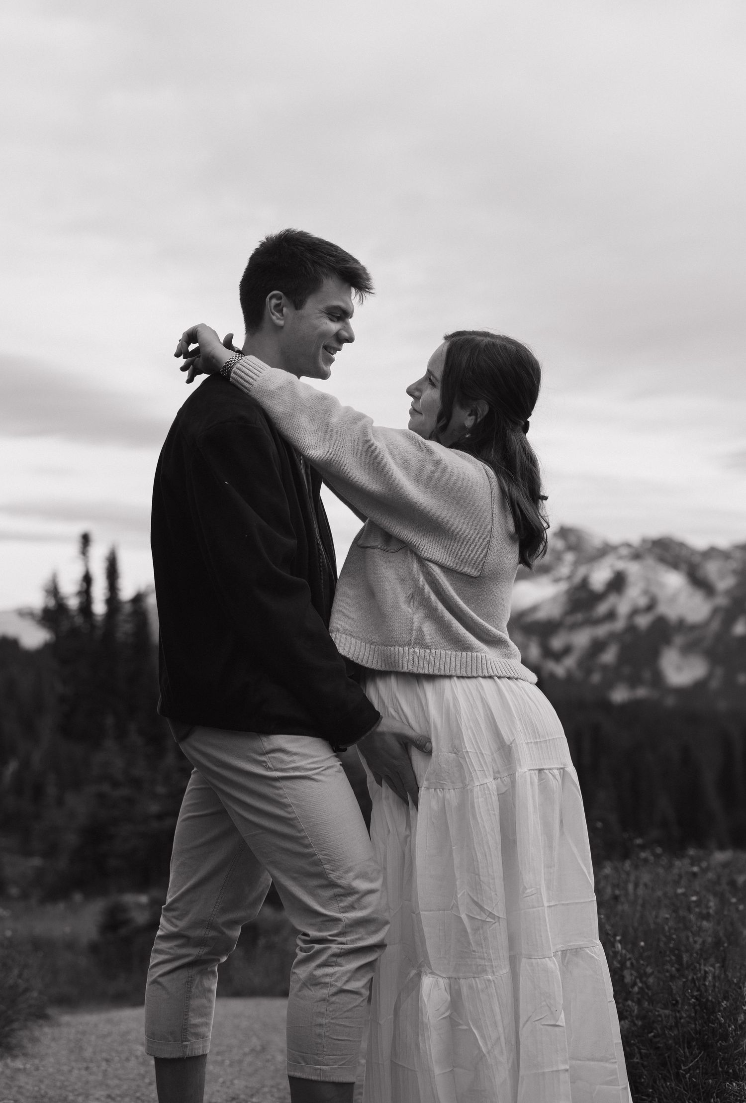 Black and white photo of a couple embracing outdoors with mountain scenery, woman in white skirt resting arm on mans shoulder.