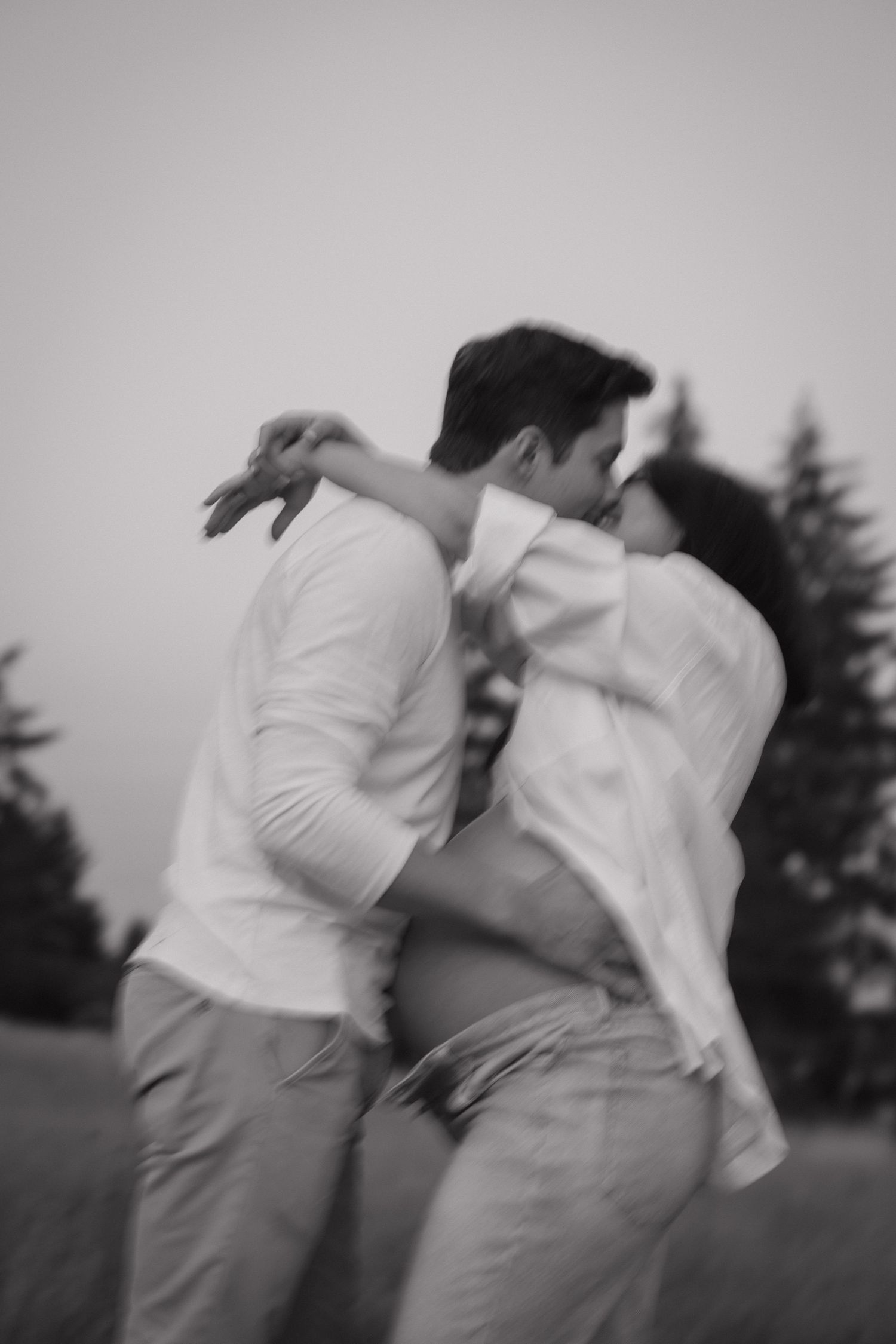 Black and white maternity photo of a couple embracing and kissing in a grassy field with evergreen trees, her bare pregnant belly showing.