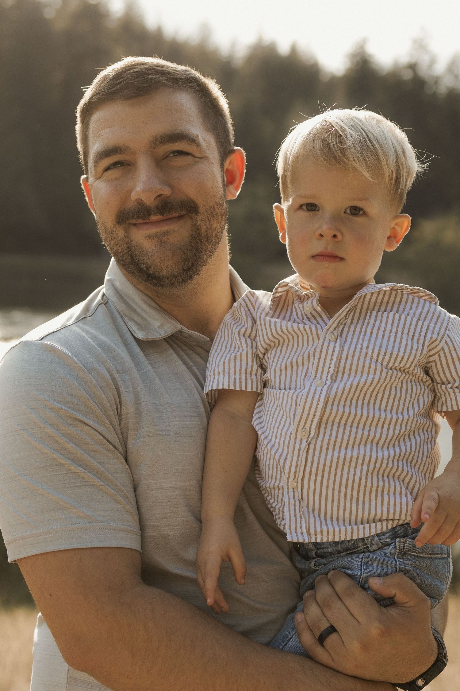 Smiling bearded father holding his young blonde toddler son outdoors in golden sunlight.