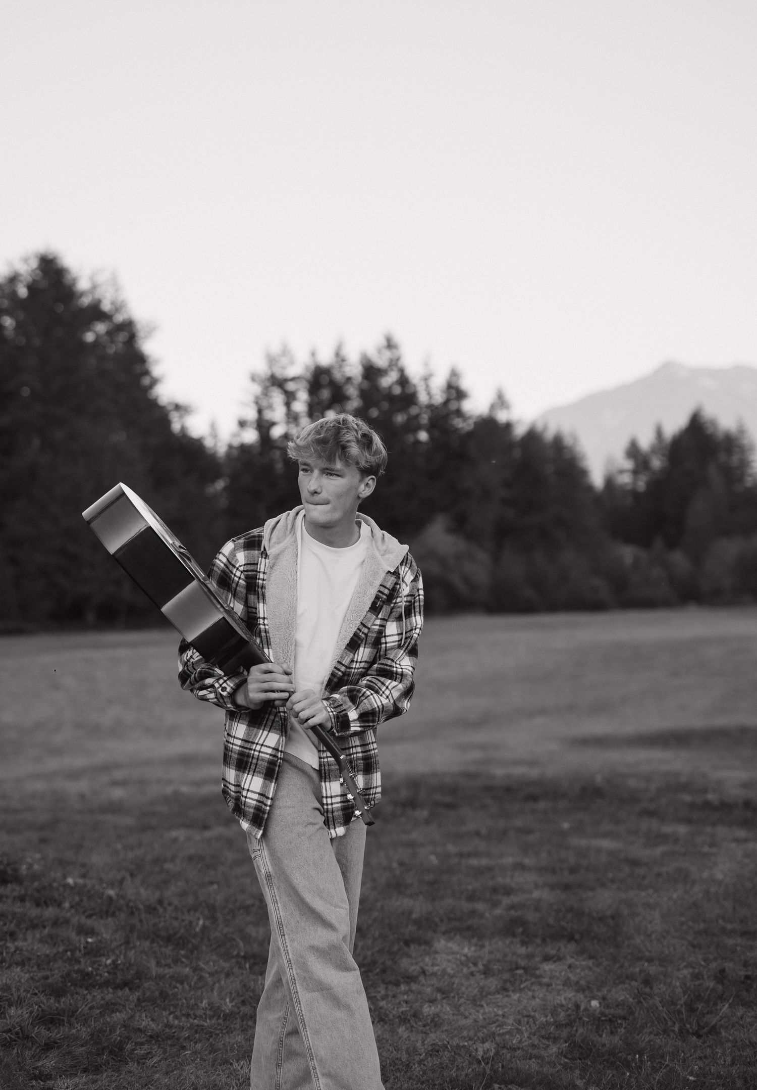 A young boy holds a guitar case in a plaid shirt outdoors in a black and white photo.