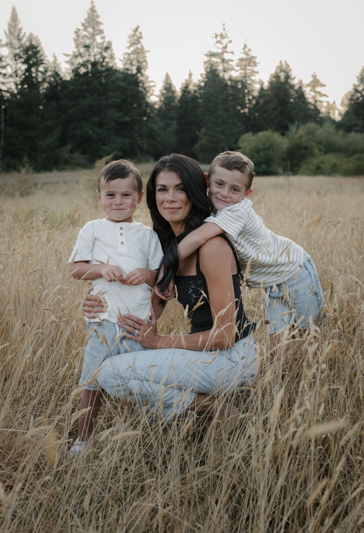 A woman with two young boys poses in a golden grass field with evergreen trees in the background.