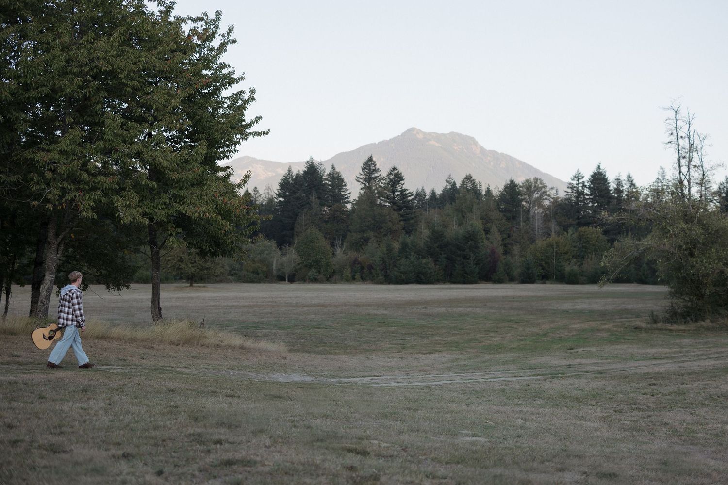 Person walking across a grassy field with tall trees and a snow-capped mountain in the background.