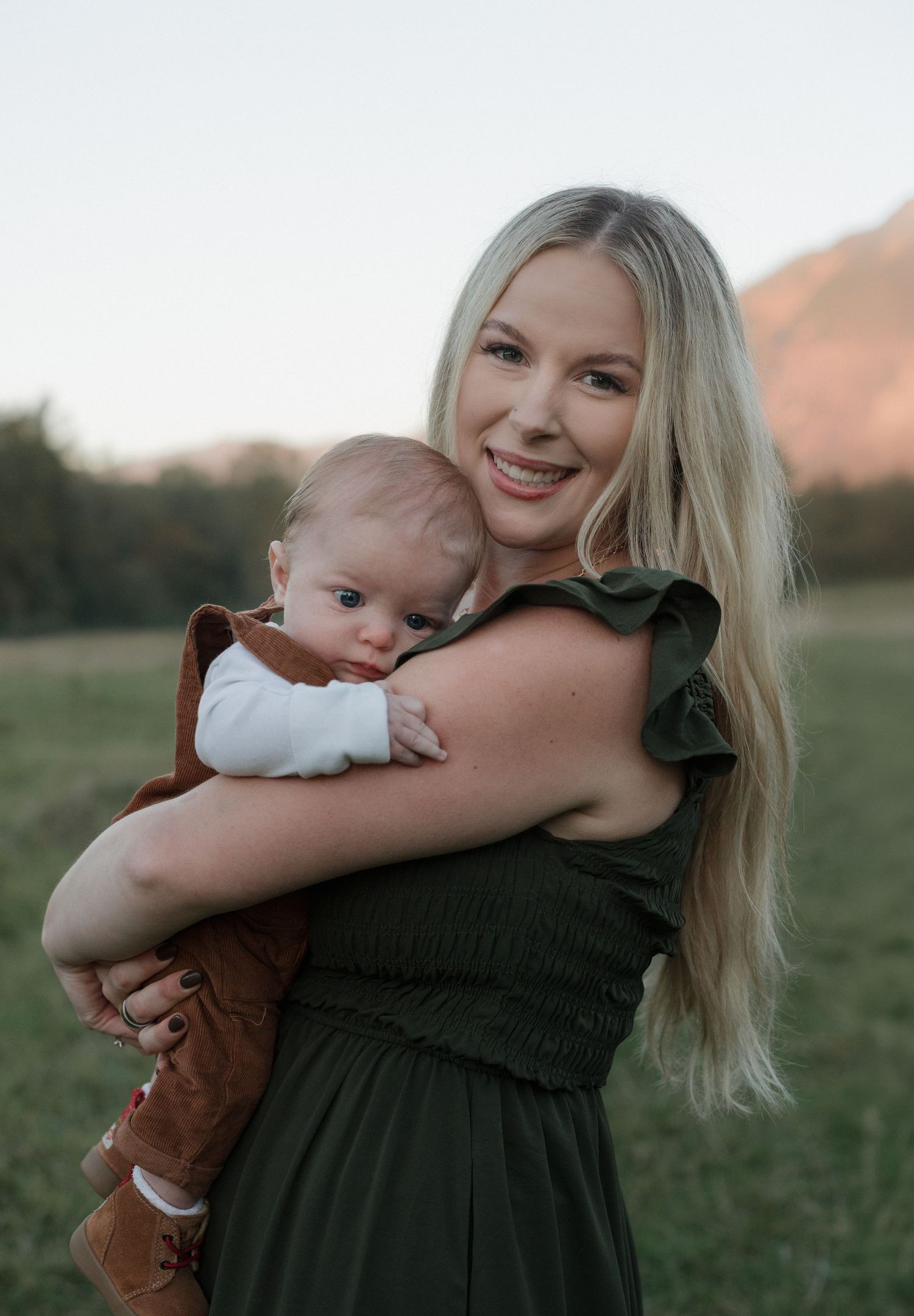 A smiling blonde woman in a green dress holds a baby in cowboy boots in a scenic outdoor field.