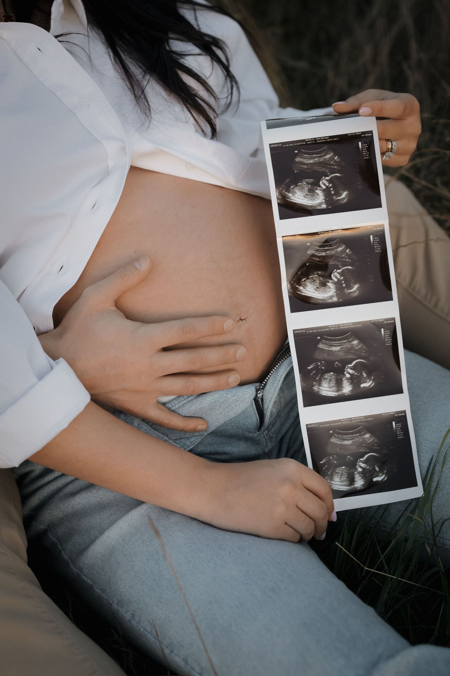 Pregnant woman in white shirt and jeans holds a strip of ultrasound photos while cradling her bare baby bump outdoors.