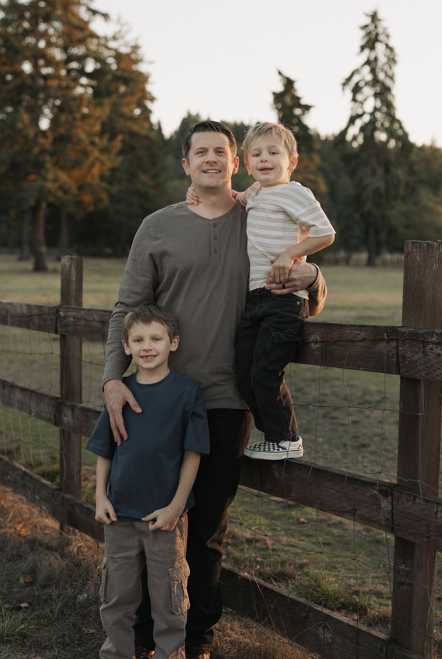 A father smiles holding his toddler on a fence while his older son stands beside them in a sunny field.