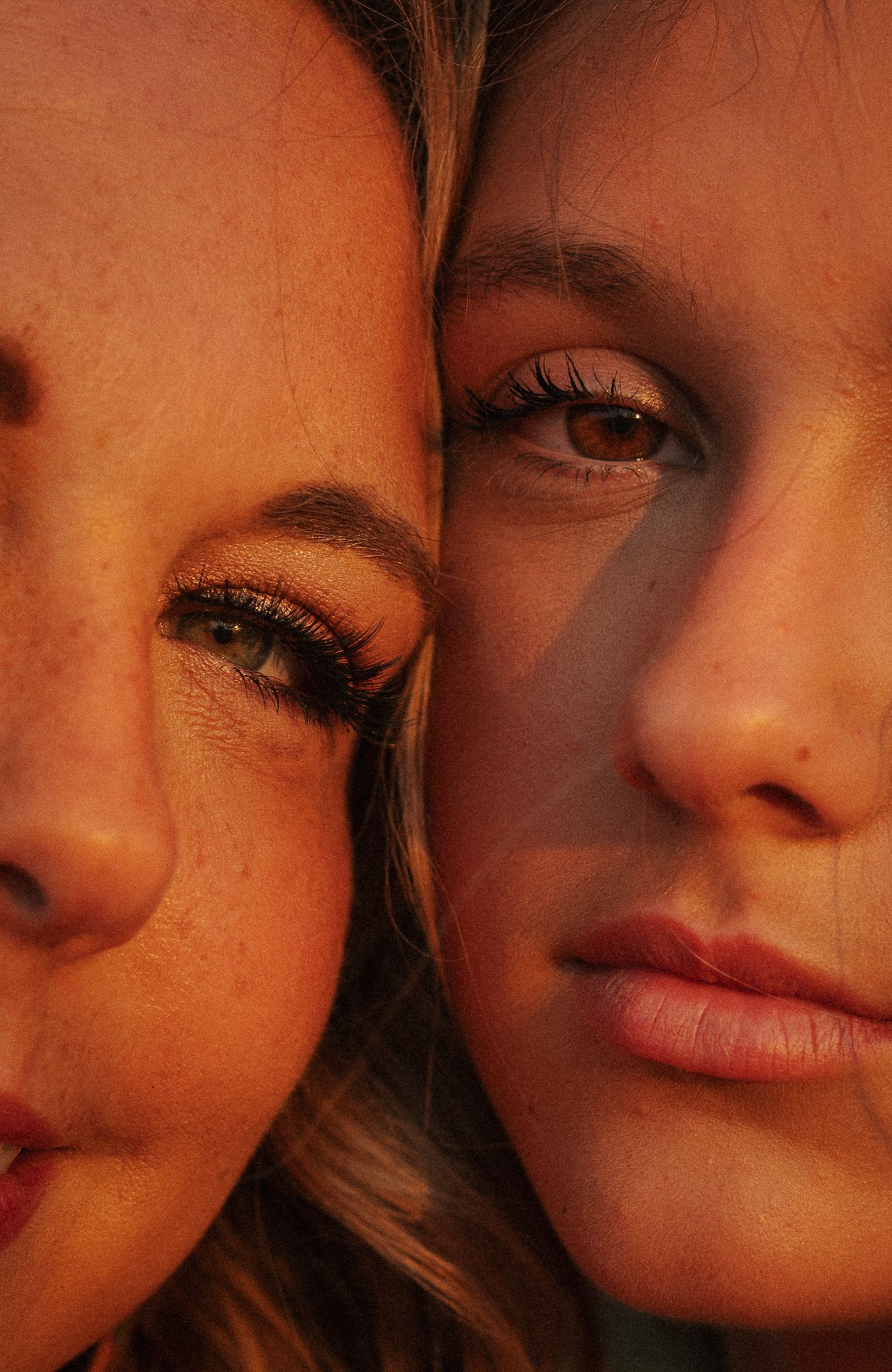 Close-up portrait of two young women with glowing skin in warm golden light, faces pressed together cheek to cheek.