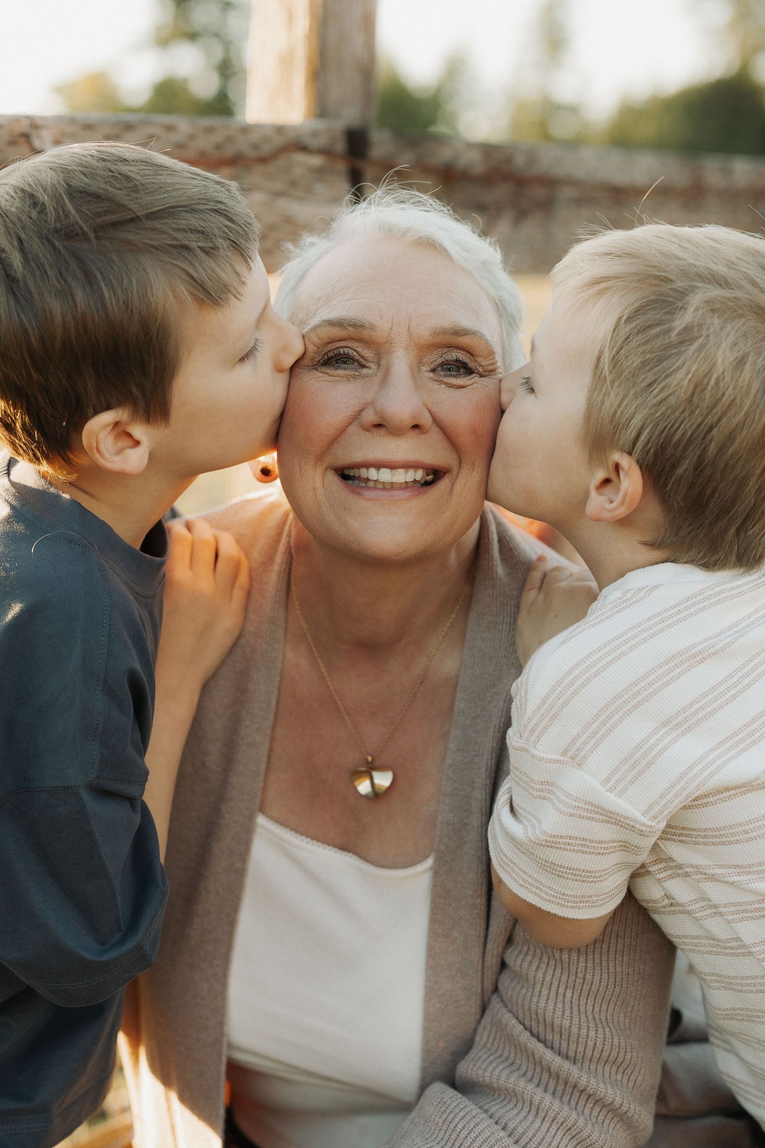 Elderly woman smiling joyfully as two young boys kiss her on each cheek outdoors in warm sunlight.