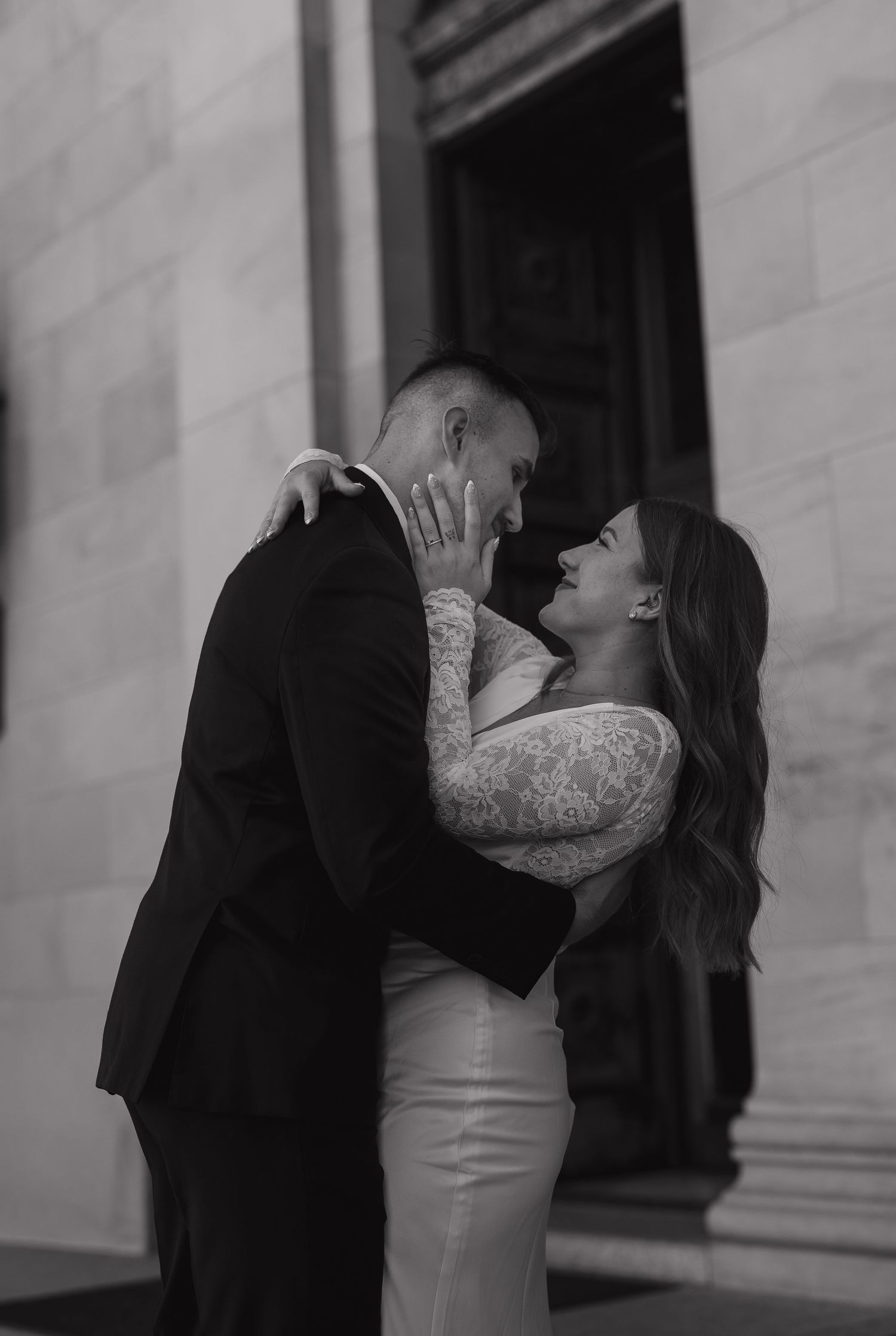 Black and white photo of a couple in wedding attire sharing a romantic moment, embracing and smiling at each other outdoors.