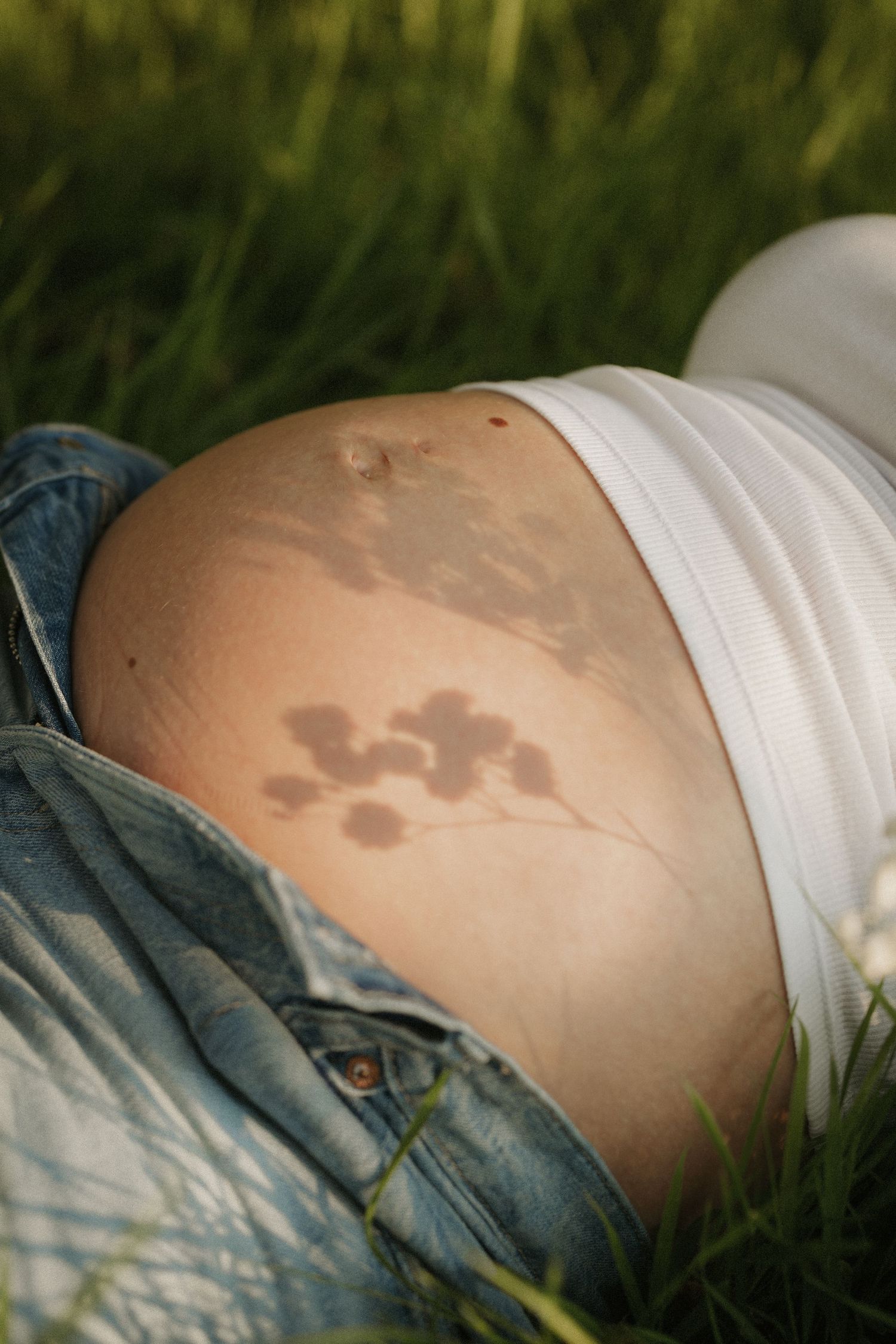 Pregnant woman lying in green grass, showing bare belly with flower tattoo, wearing white top and open denim jeans.