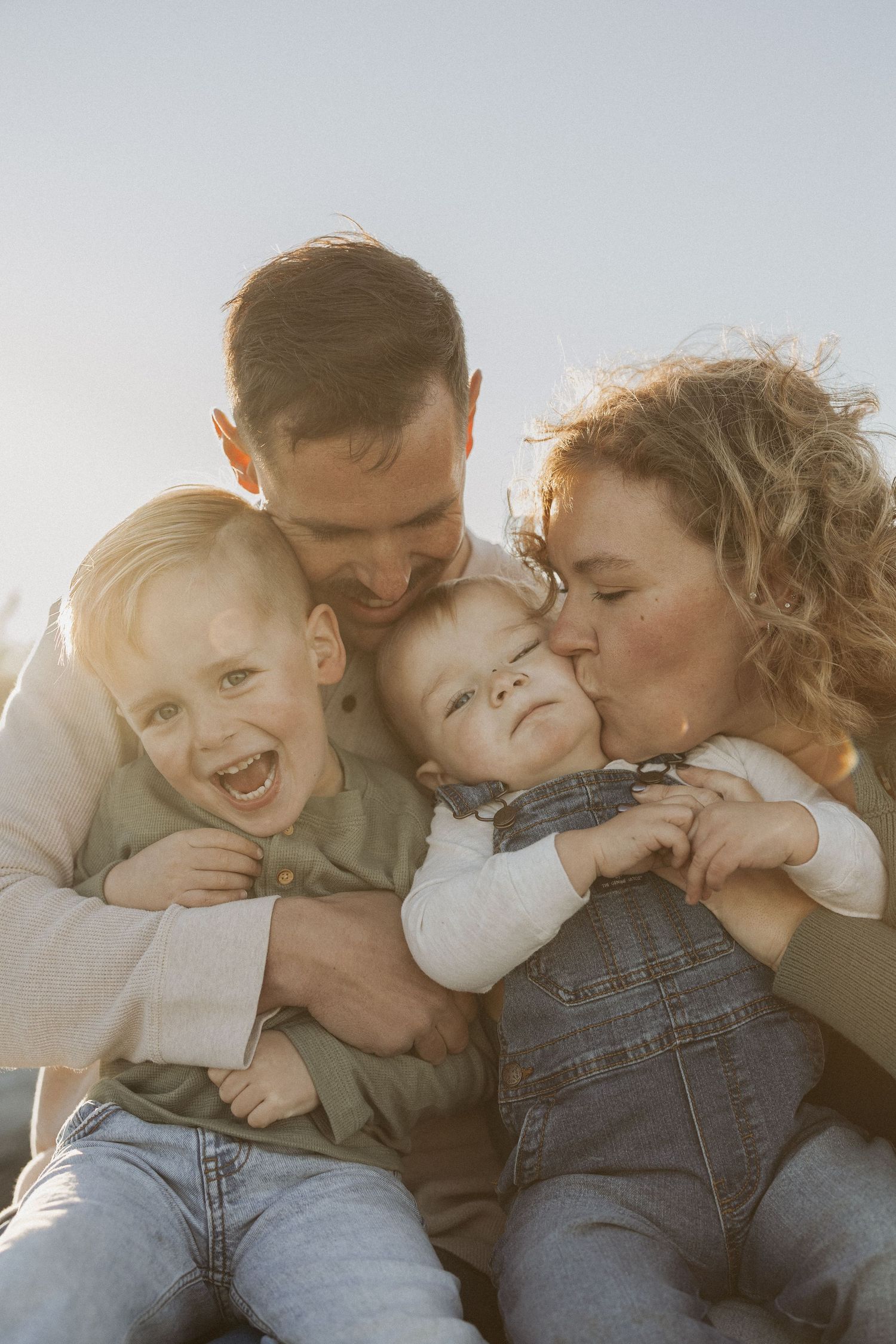 Happy family with two young children sharing kisses and laughter outdoors in warm golden sunlight.