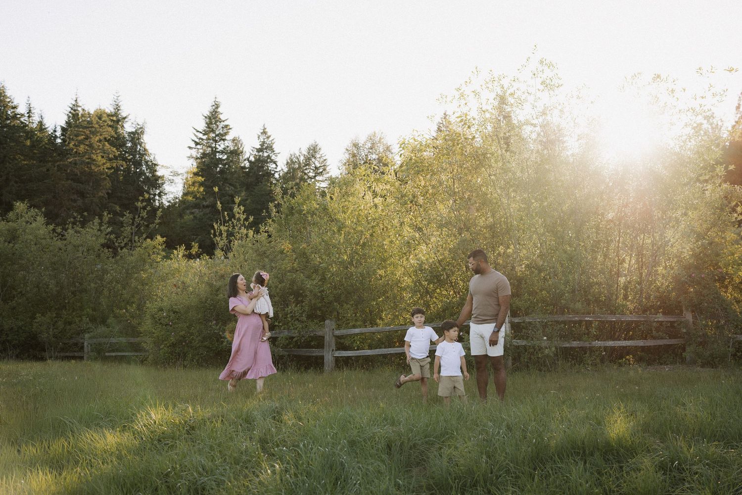 Family of four walking in a sunlit green meadow with trees and wooden fence in background.
