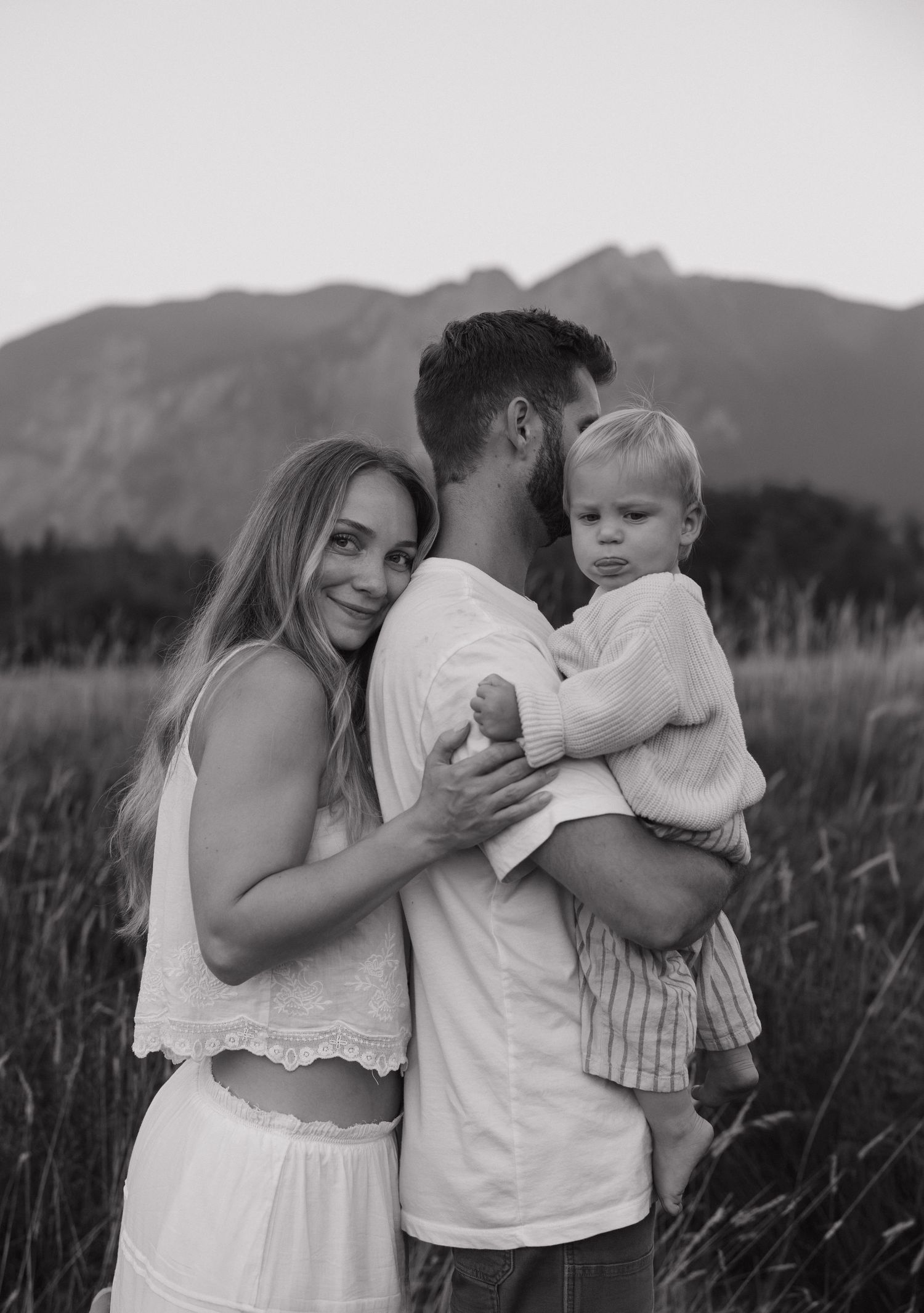 Black and white family portrait of a couple with their toddler in a field with mountains behind them.
