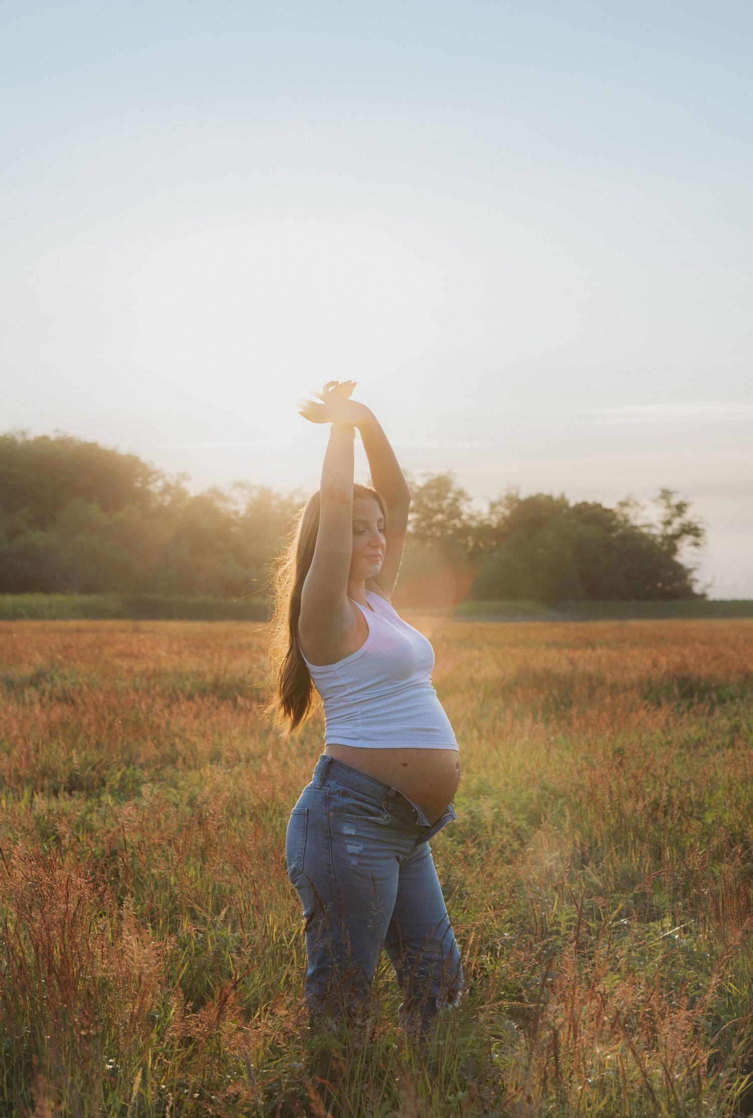 Pregnant woman in white tank top and jeans poses in sunlit field at golden hour with arms raised.