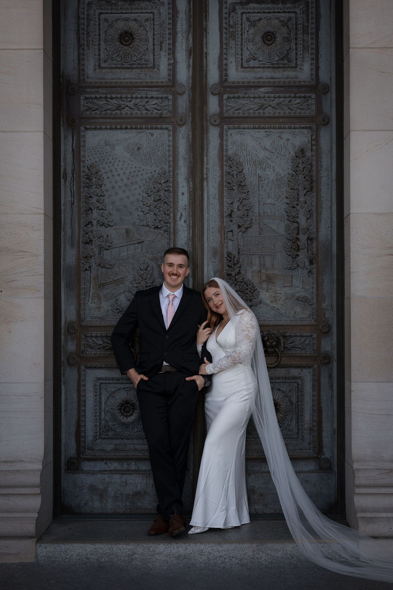Bride in white gown and veil with groom in black suit posing before ornate bronze doors.