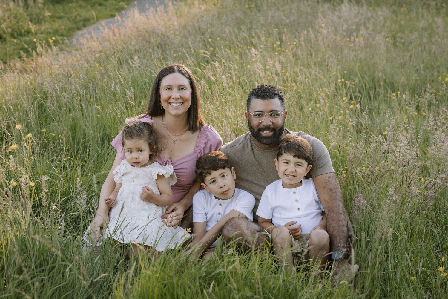Happy family of five sitting together in tall golden grass smiling at camera.