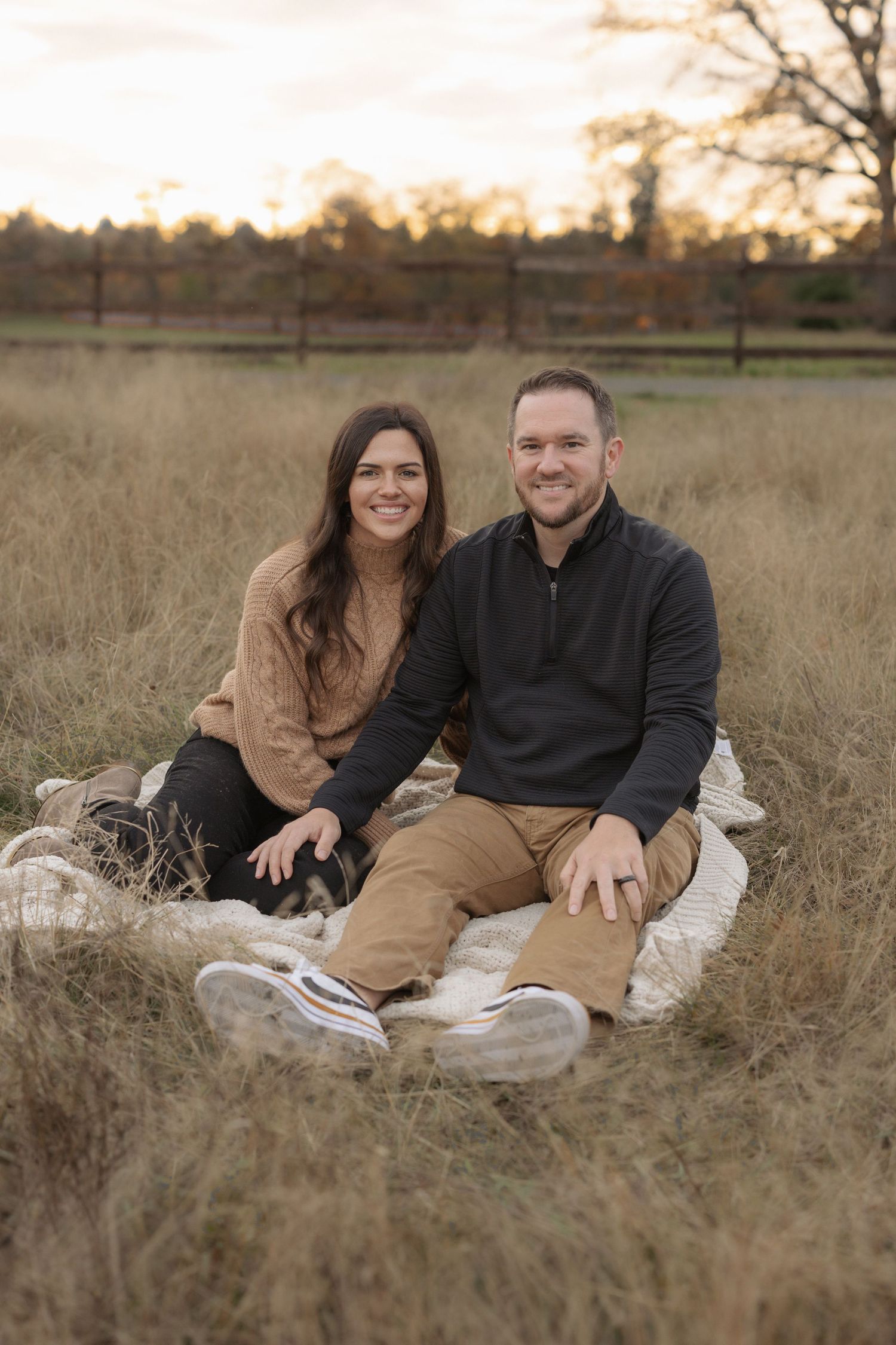 Couple sitting on blanket in golden field at sunset, woman in tan sweater and man in black pullover smiling.