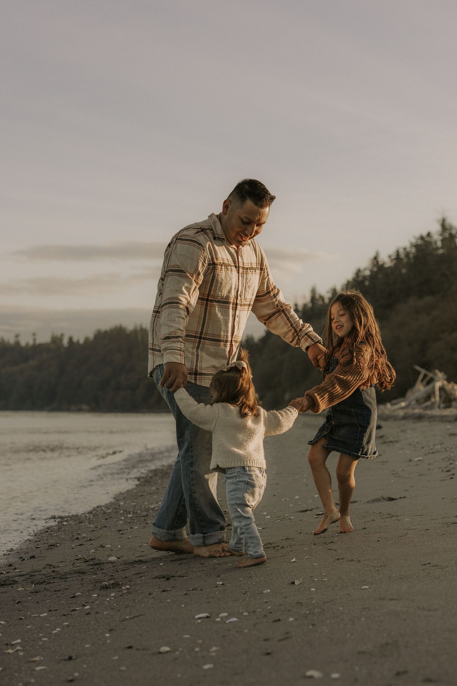 Father dancing with two young daughters on a scenic beach at golden hour, trees and calm water in the background.