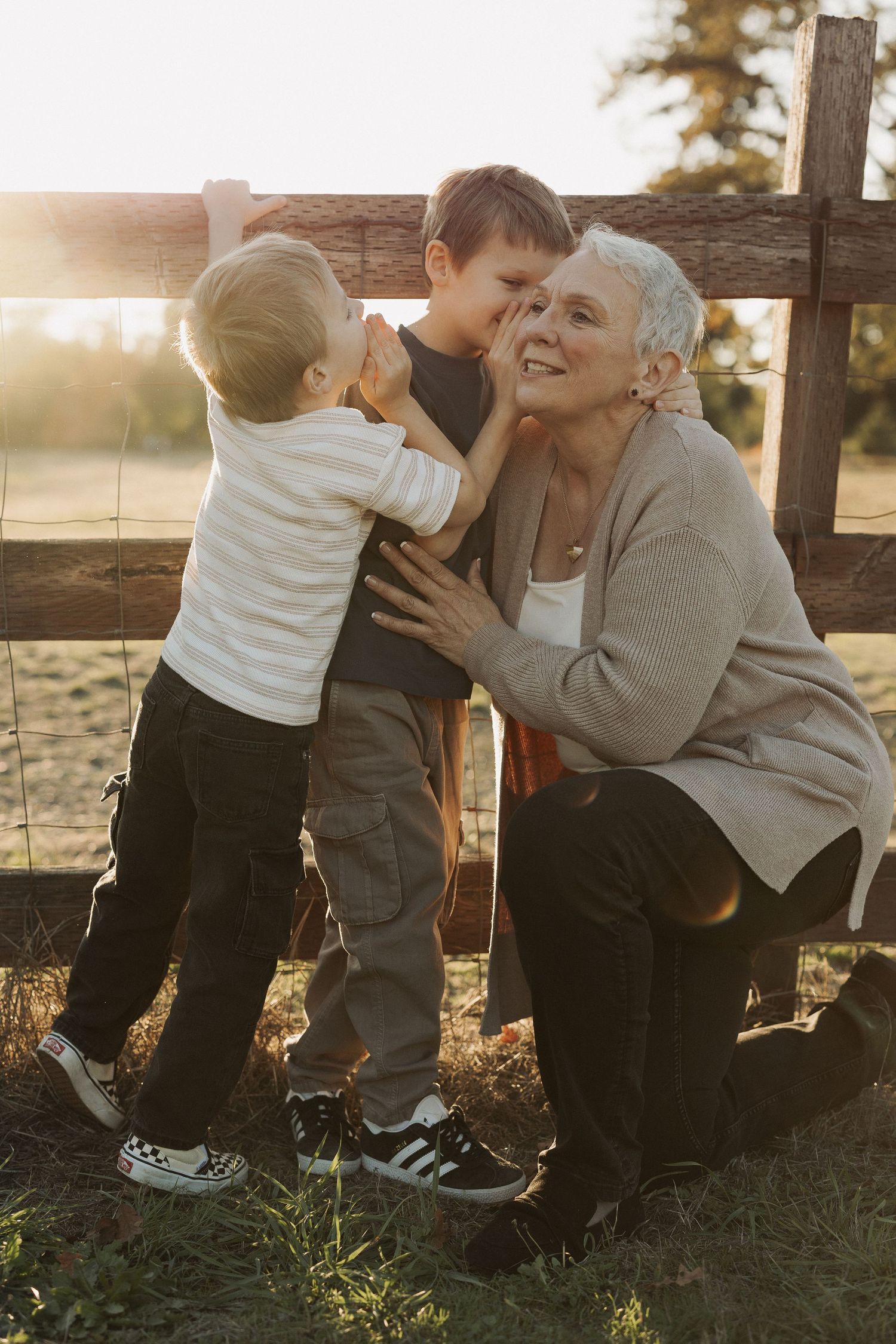 Grandmother kneeling by a wooden fence at sunset, sharing a joyful moment with two young grandsons outdoors.