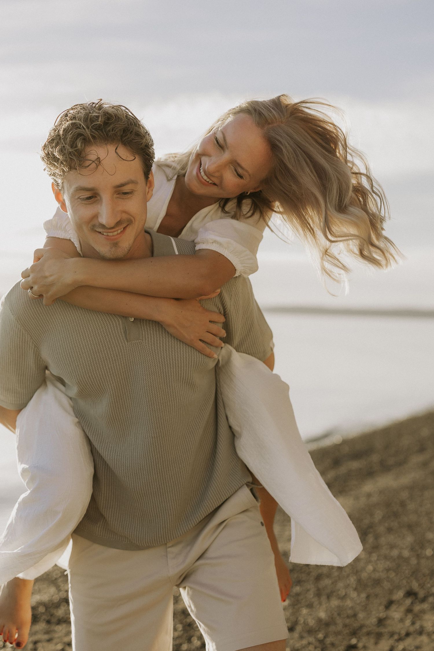 A smiling man gives a laughing woman a piggyback ride on a rocky beach, both dressed in casual neutral tones.