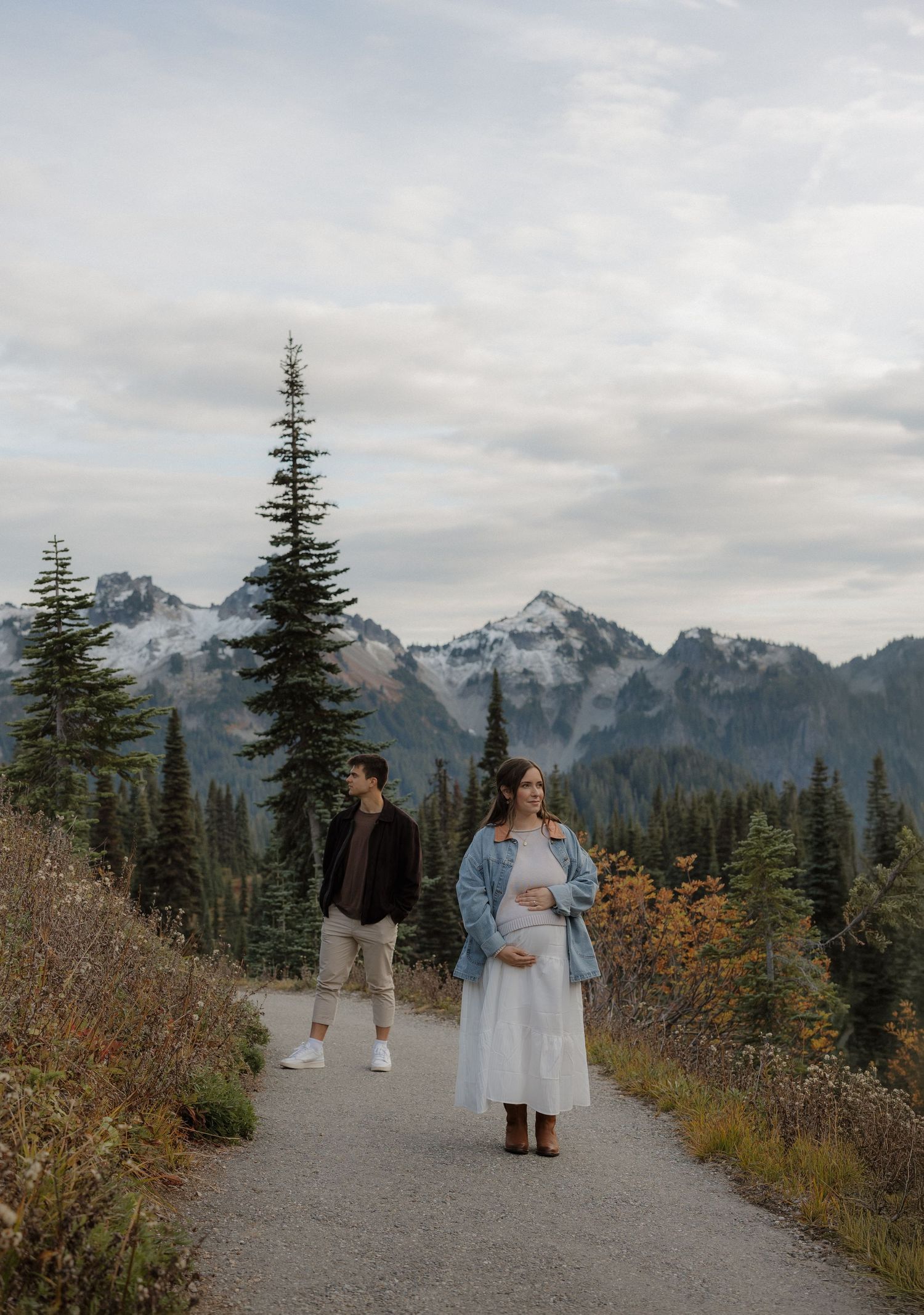 Couple on mountain trail, pregnant woman in white dress and denim jacket, snow-capped peaks and evergreens behind them.