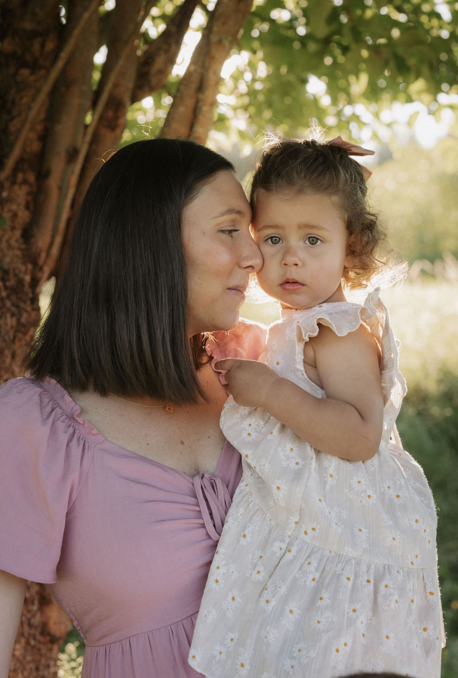 A mother in a pink dress tenderly holds her toddler daughter in a white floral dress outdoors under a tree.