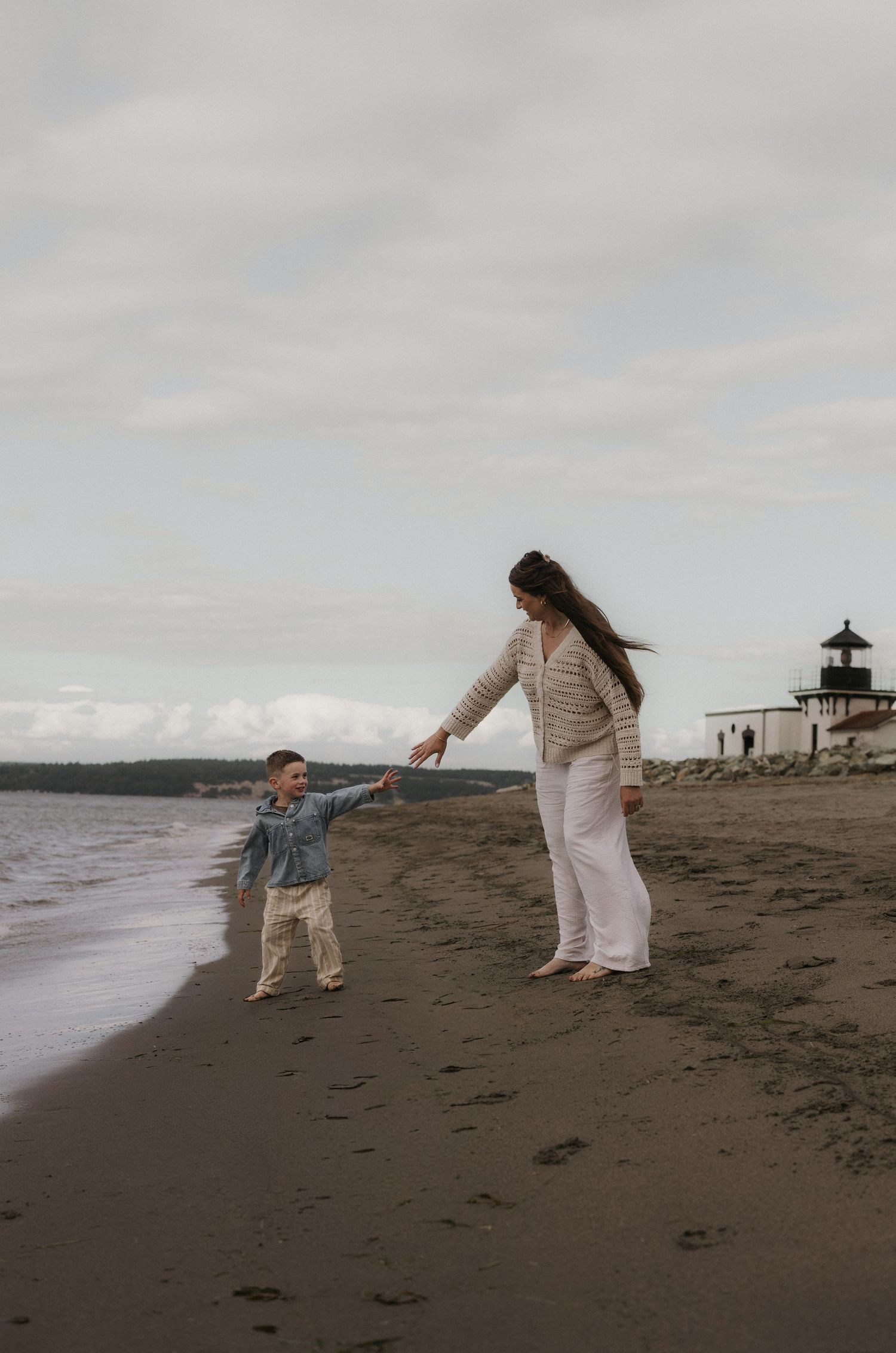 A mother and young child hold hands while walking along a sandy beach near a lighthouse on a cloudy day.
