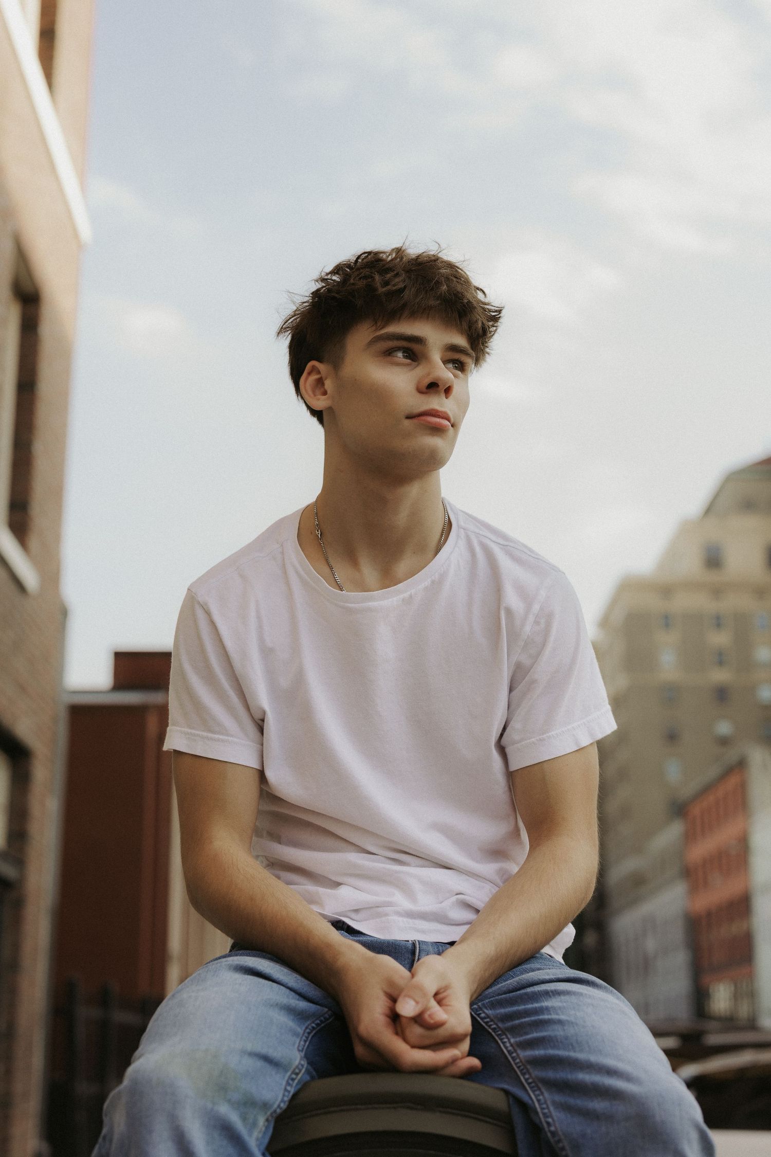 Young man in white t-shirt and jeans sitting outdoors in urban setting, looking thoughtfully to the side.
