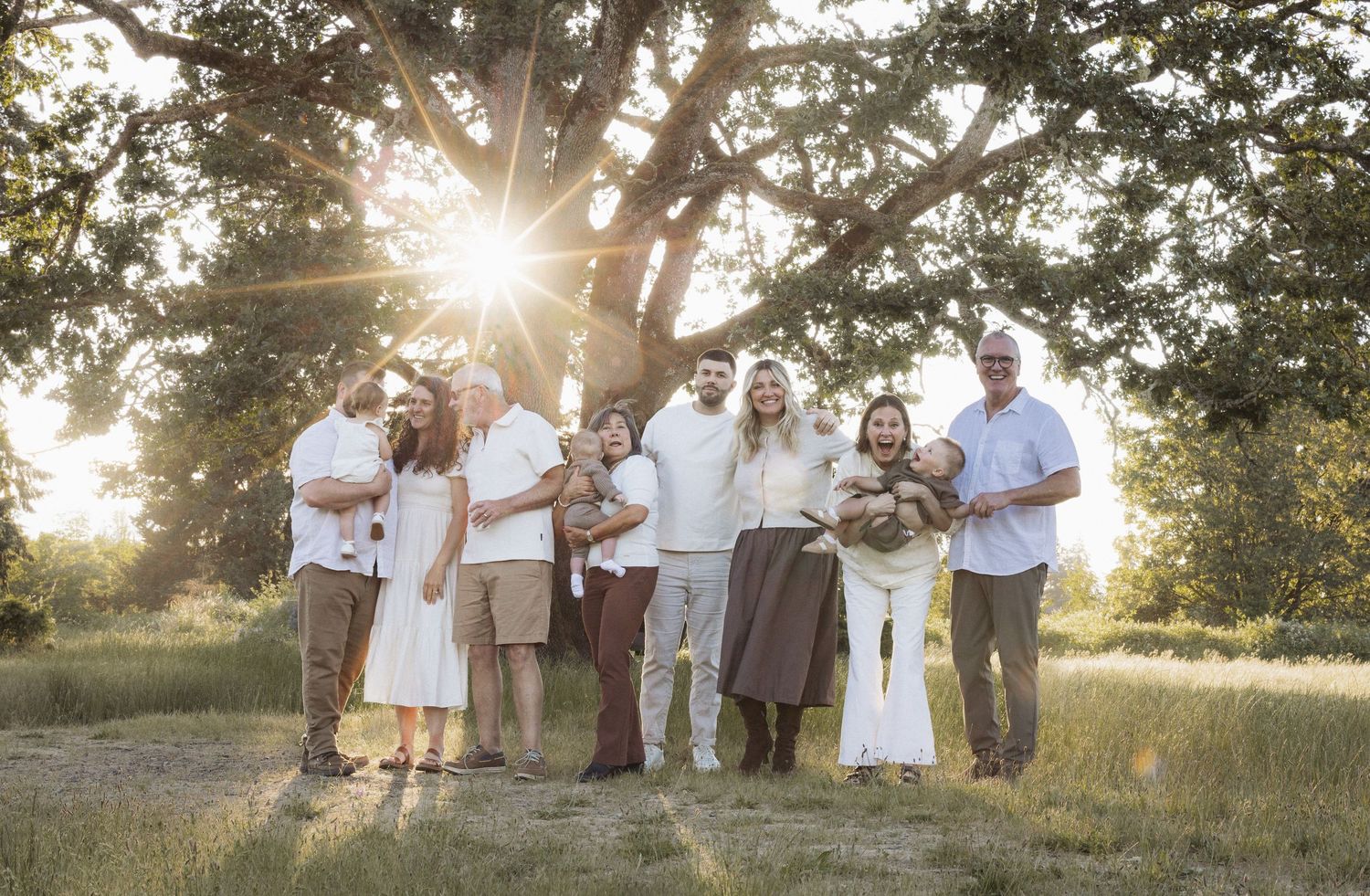 Family portrait of multigenerational group posing outdoors under large oak tree with golden sunset light filtering through branches.