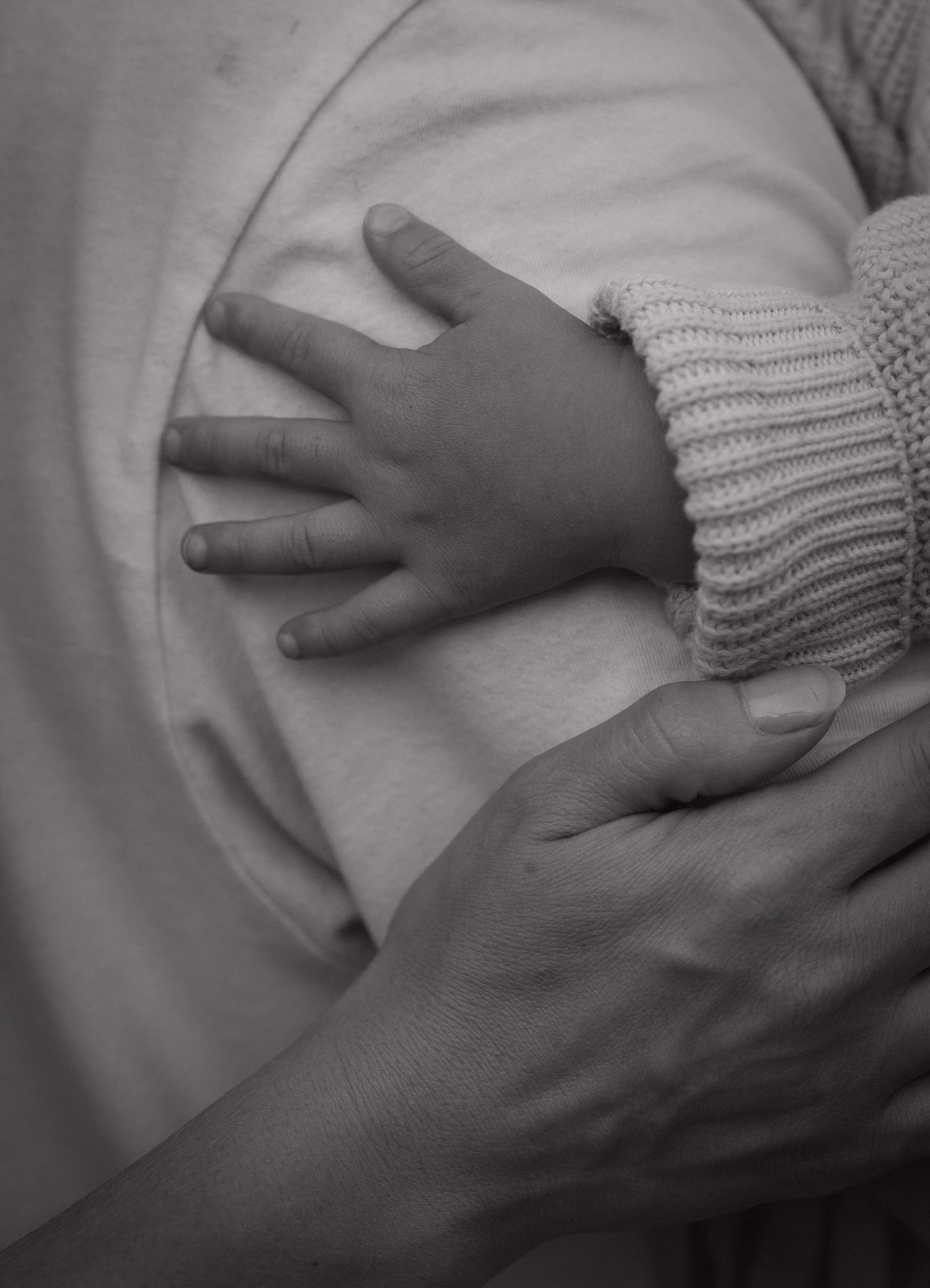 Black and white photo of a baby's tiny hand resting on an adult's hand, wearing a knit sweater sleeve.
