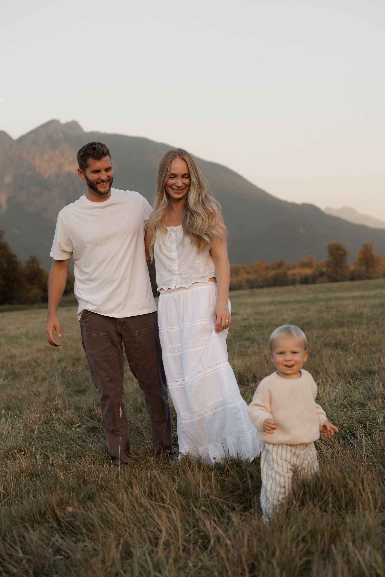 A happy family of three walks through a mountain meadow at sunset, parents smiling beside their toddler.