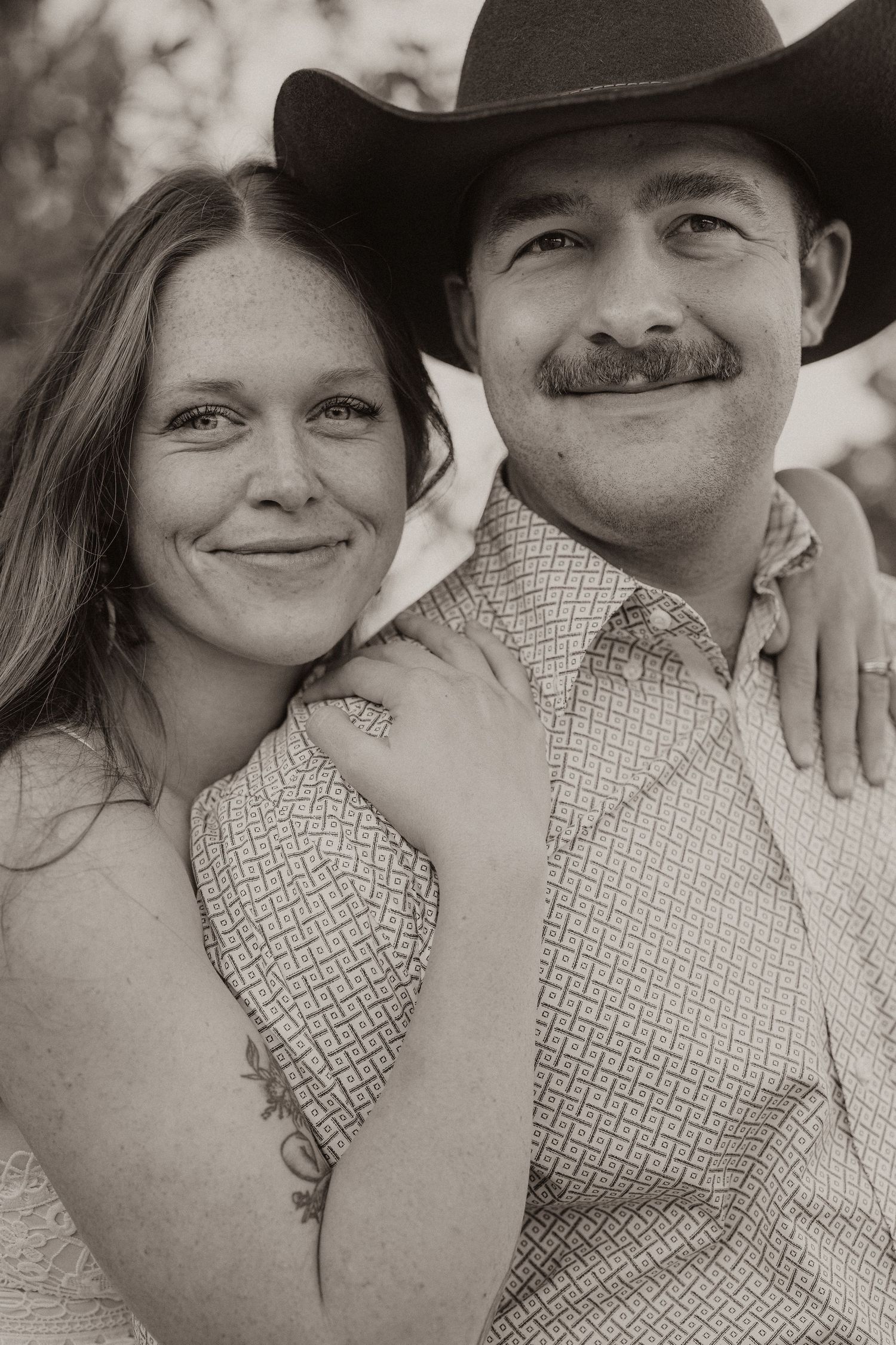 Sepia-toned portrait of a smiling couple, man with mustache and cowboy hat, woman with tattoo embracing him.
