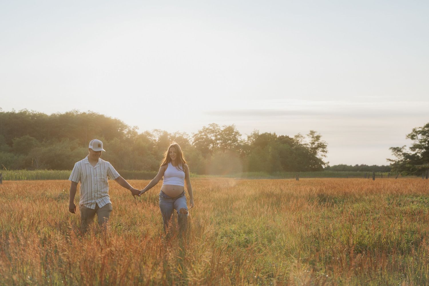 Couple holding hands walking through golden field at sunset with trees in background.